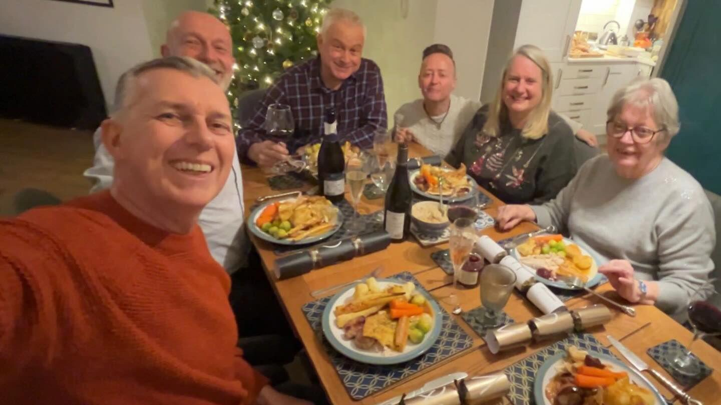 Six people, four men and two women smiling for a selfie at a table. They are eating Christmas lunch with crackers between them. There is a lit up Christmas tree behind them