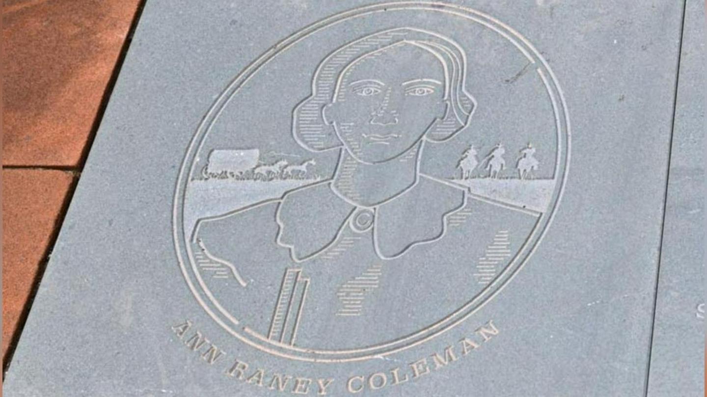 An image of Ann Raney Coleman etched on to a flagstone. The design shows a head and shoulders image of a woman with short hair. Behind her are three men on horses and a horse-drawn cart.