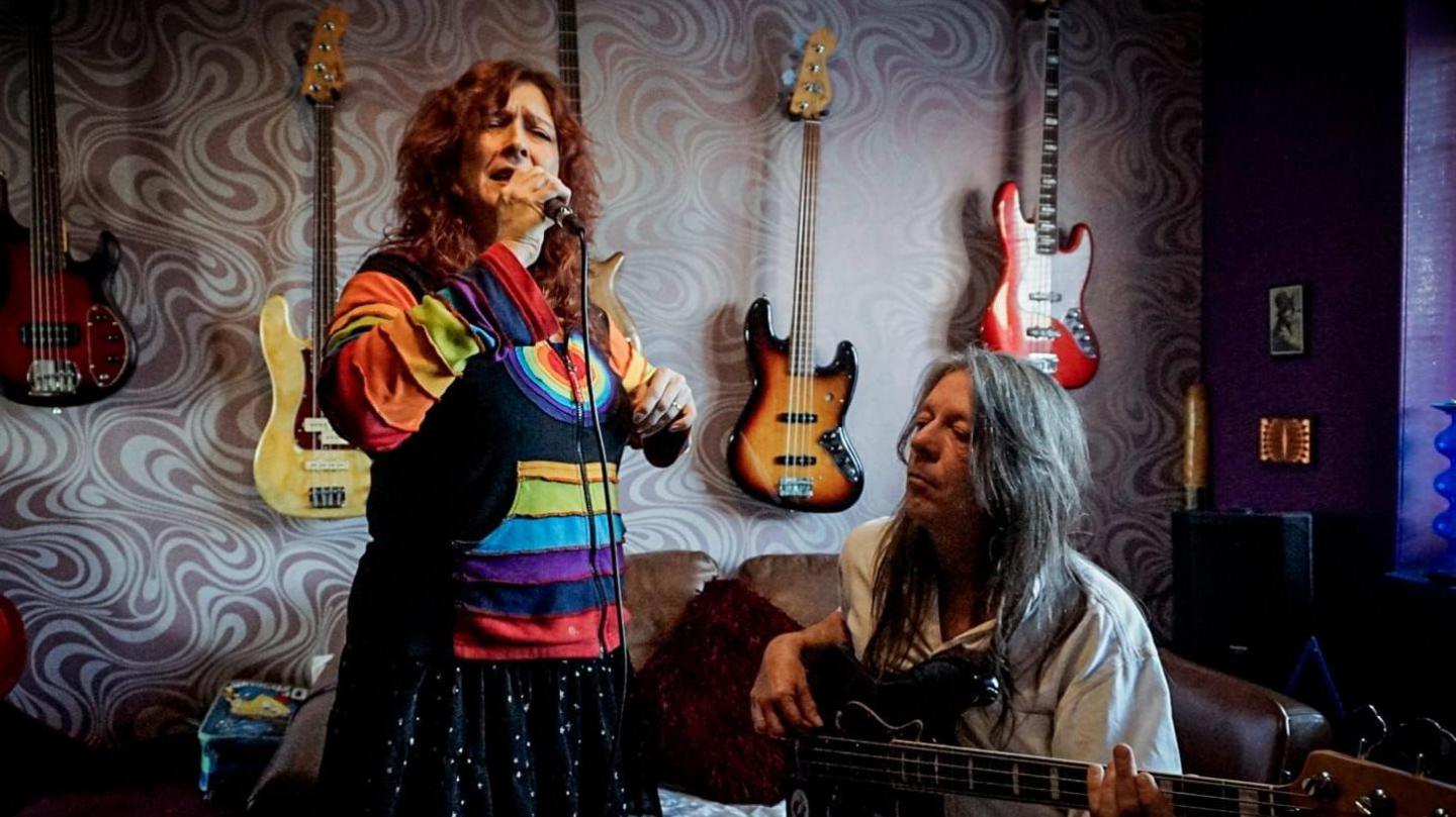 A woman with reddish-brown medium-length hair sings passionately into a microphone with her eyes closed. She wears a colourful, rainbow-striped cardigan and stands beside a seated man with long grey hair, who is playing bass guitar. The wall behind them is adorned with electric guitars in different colours.