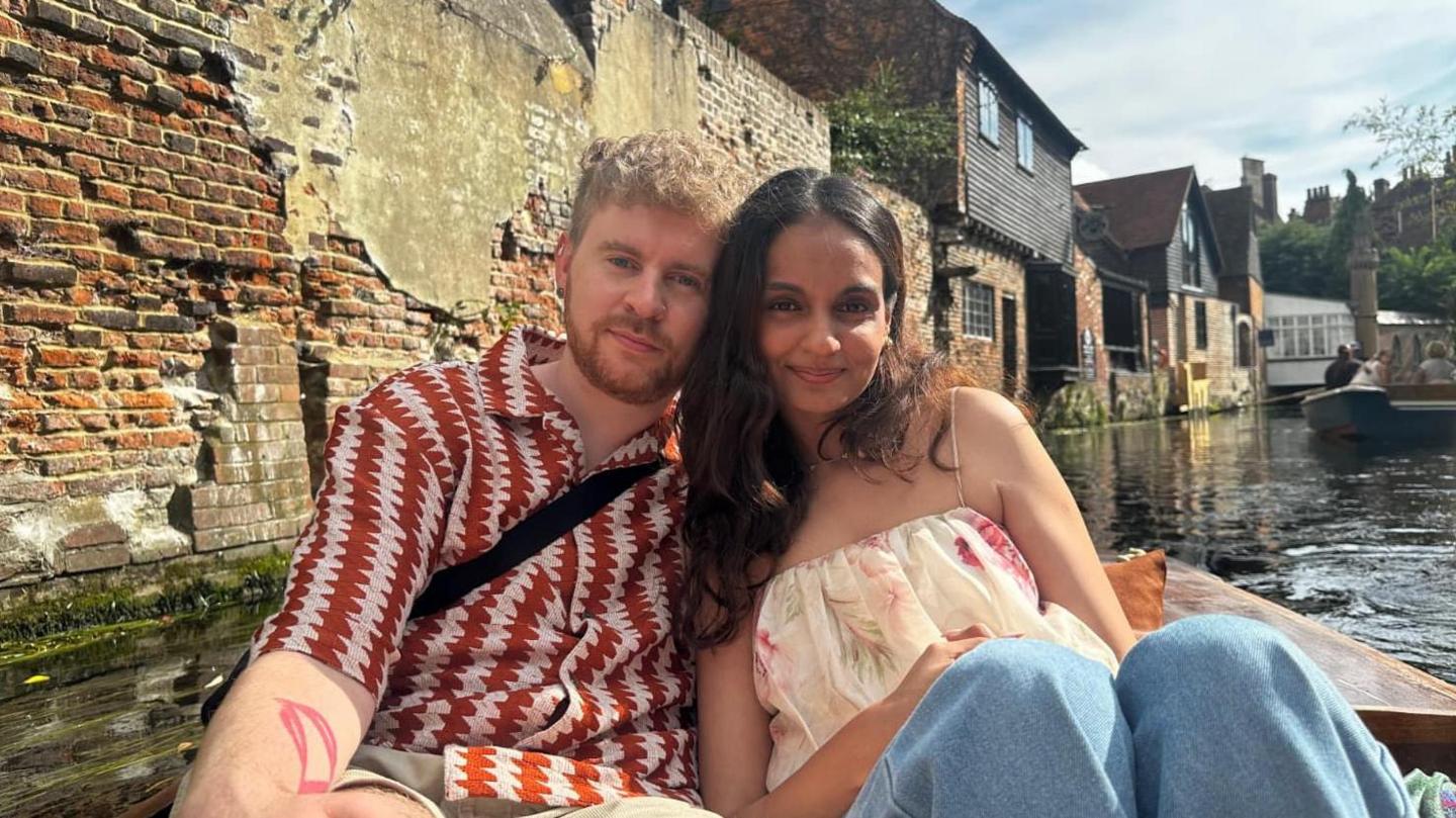 Conor Kehoe and Manisha Dev are sitting in a punt on a river smiling at the camera. He has wavy fair hair, a beard and moustache and is wearing a red and white patterned shirt. She has long, wavy dark hair and is wearing a floral top and jeans