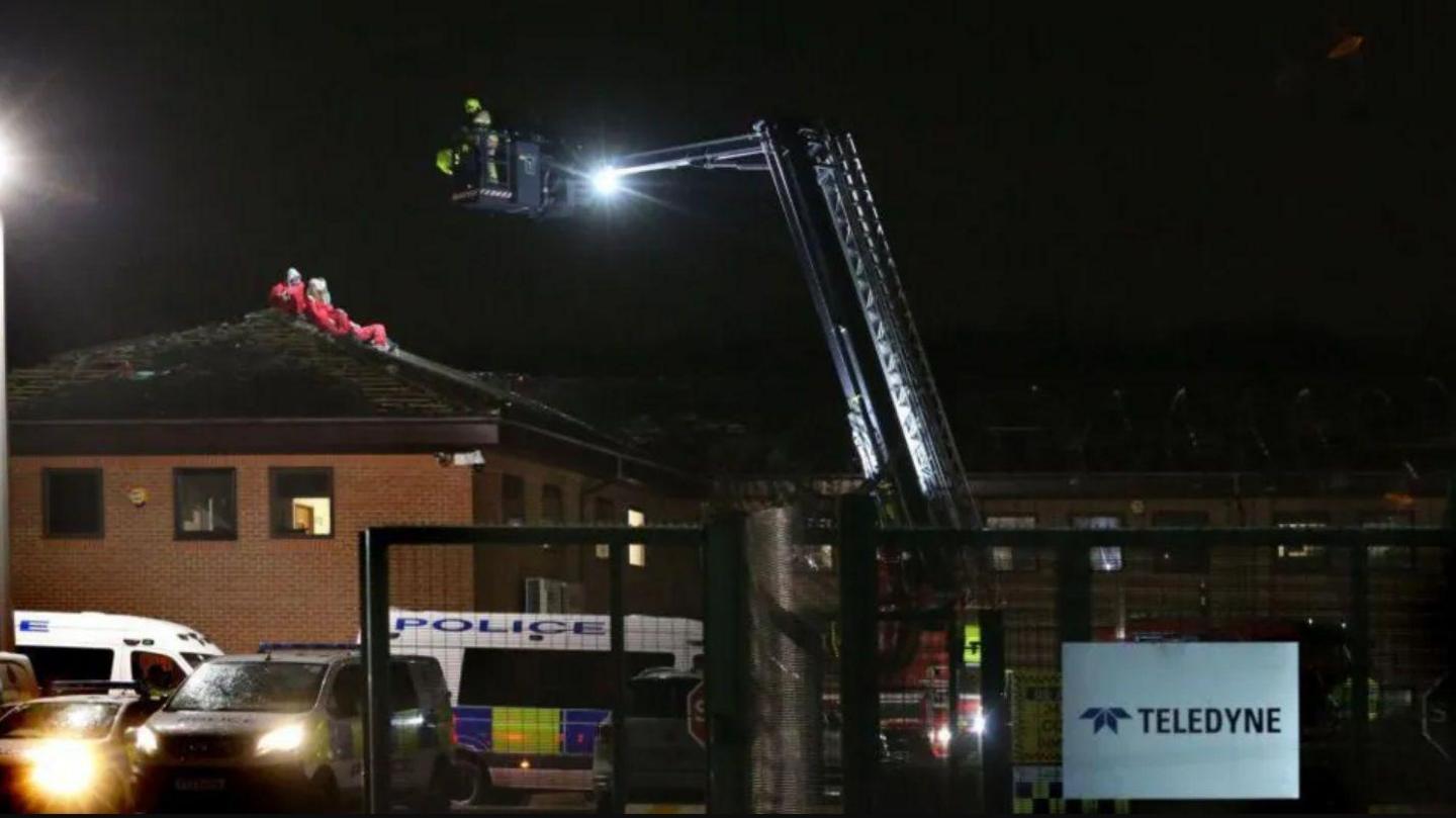 A cherry picker with two people on it hovers above the roof. People in red suits can be seen lying or sitting on the roof. Police vans are parked below and a sign that reads "Teledyne" is fixed to the fence on the right.