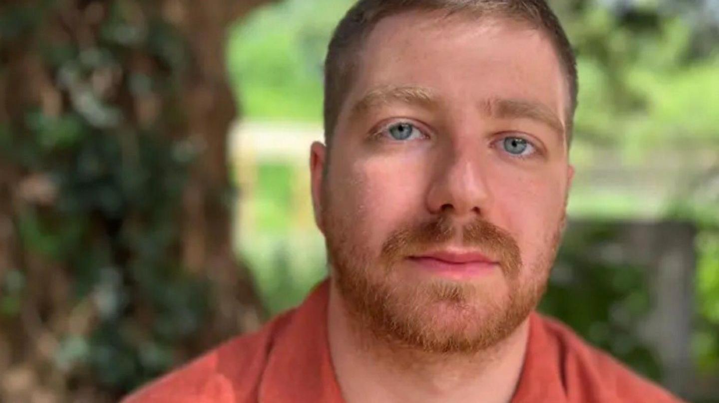 Paddy wearing an orange button-down shirt, looking at the camera. He appears to be sitting outside, with greenery behind him.