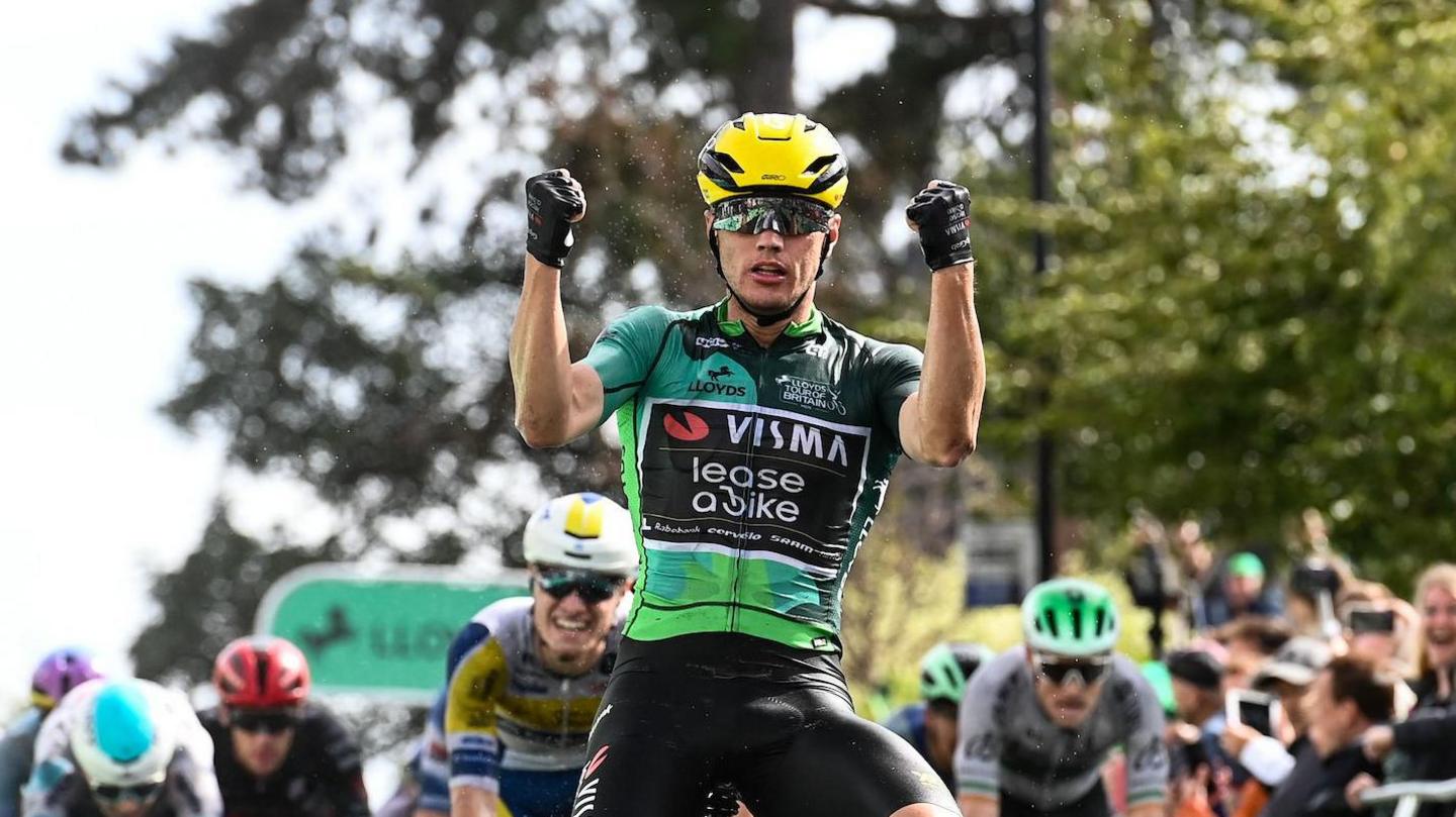 Olav Kooij raises his fists in the air as he cycles over the finish line. Other cyclists can be seen behind him. Spectators watch on from the side of the road. 