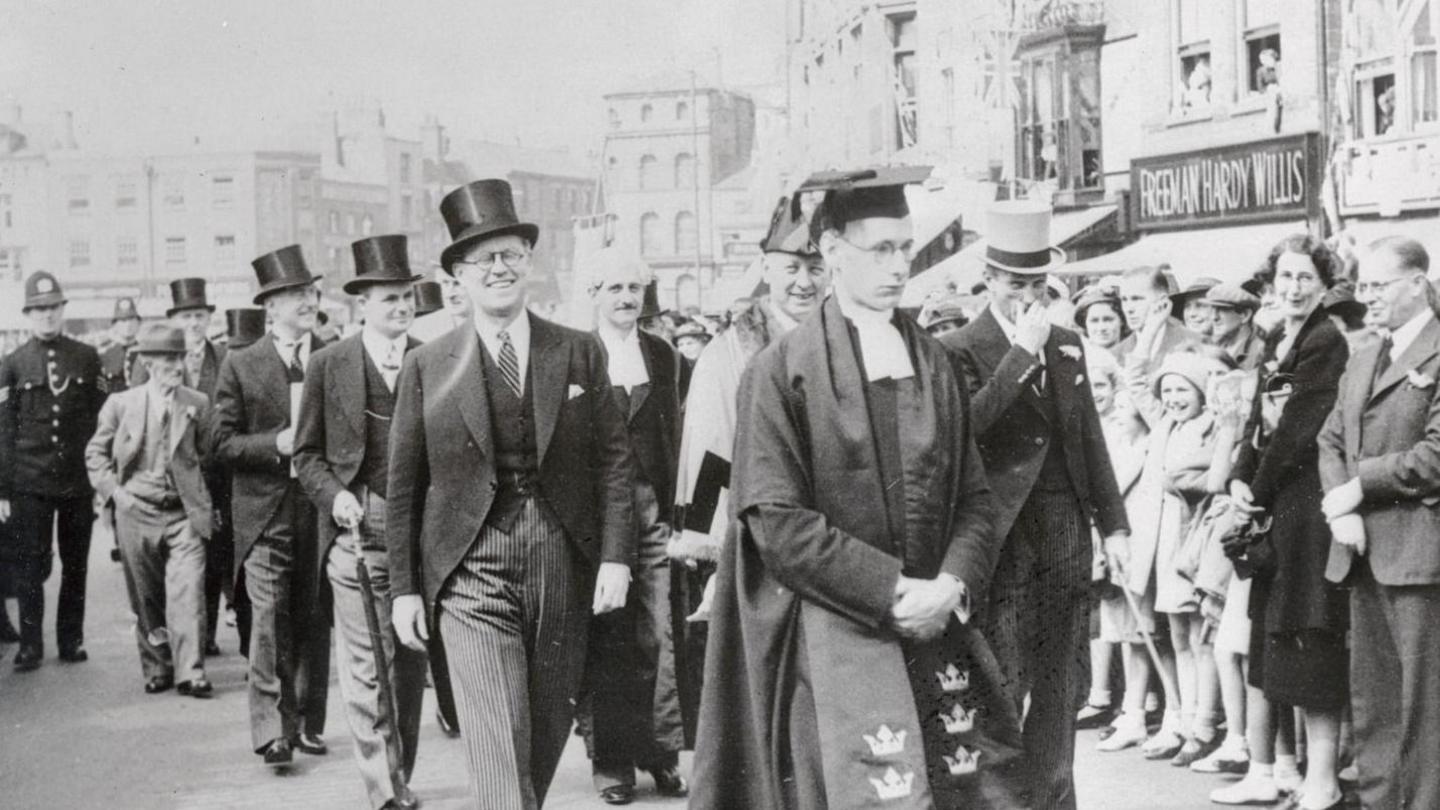 A black and white image showing the centre of Boston, UK. A civic party is processing through the town. The men are wearing morning suits and top hats. Joseph Kennedy is pictured in the first row of the procession. Crowds stand to the side looking on.