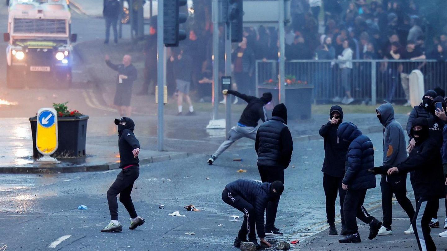 A group of about nine young people dressed in dark clothing with their faces covered on a road.
They are throwing bricks and bottles.
There is a bigger crowd in the background looking on and a police vehicle.