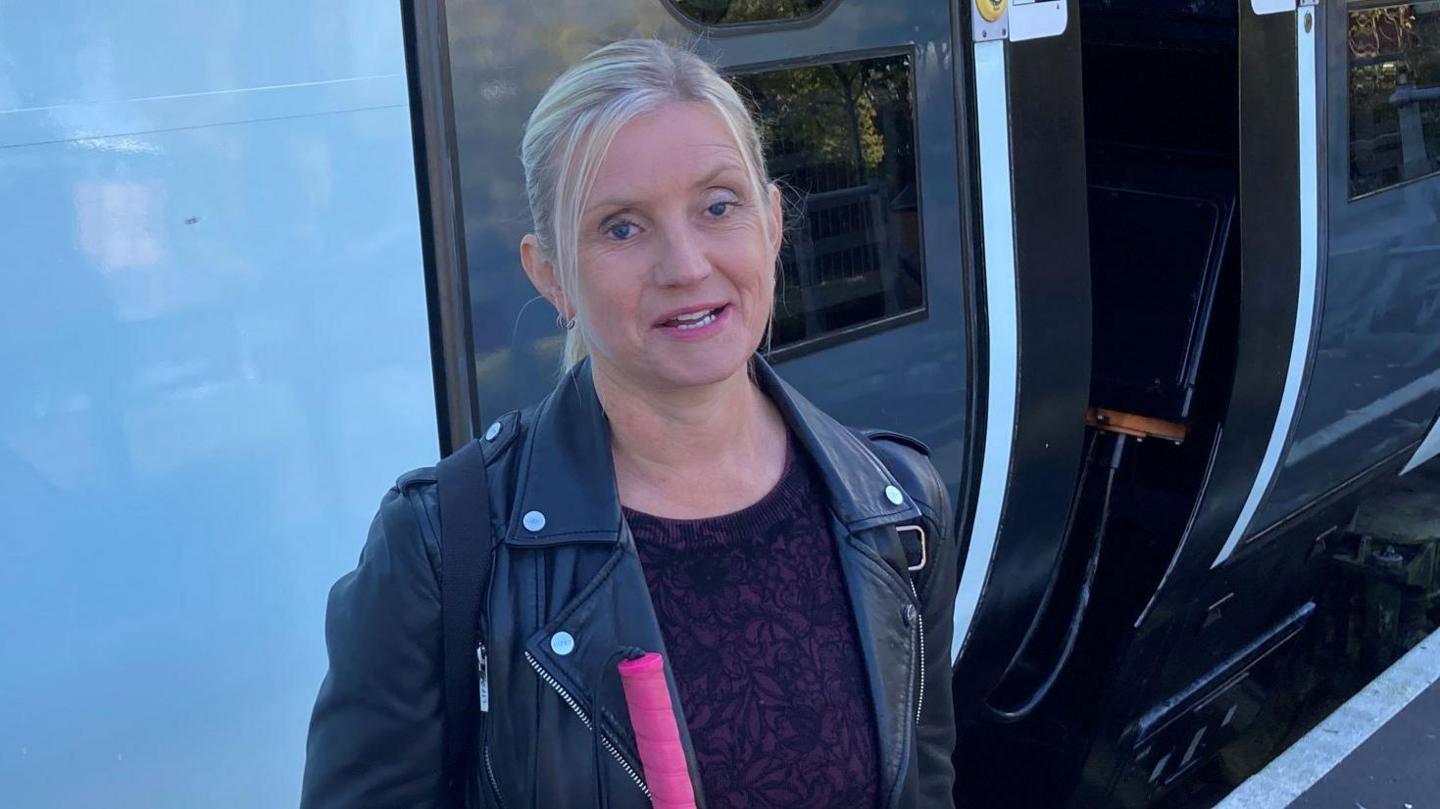 Kelly Barton has blonde hair tied back in a ponytail. She is wearing a black leather jacket and is standing next to a train carriage.