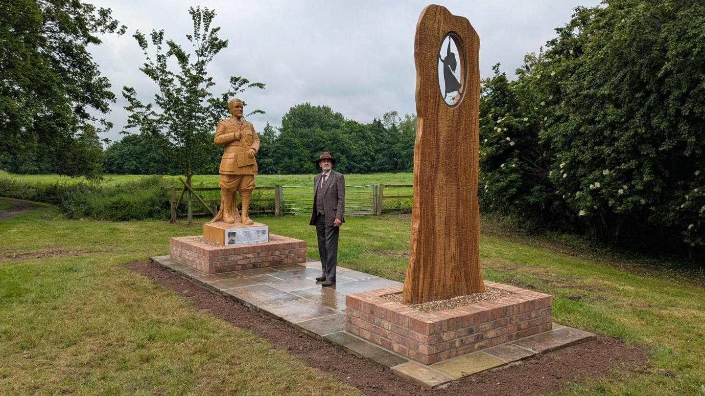 A man wearing a trilby hat and brown three-piece suit is standing on paving stones in between two wooden sculptures, erected either side of him. One of the sculptures is a man holding a book, while the other has a small circle cut out with a silhouette of a woman dancing inside. The sculptures are in grassland.