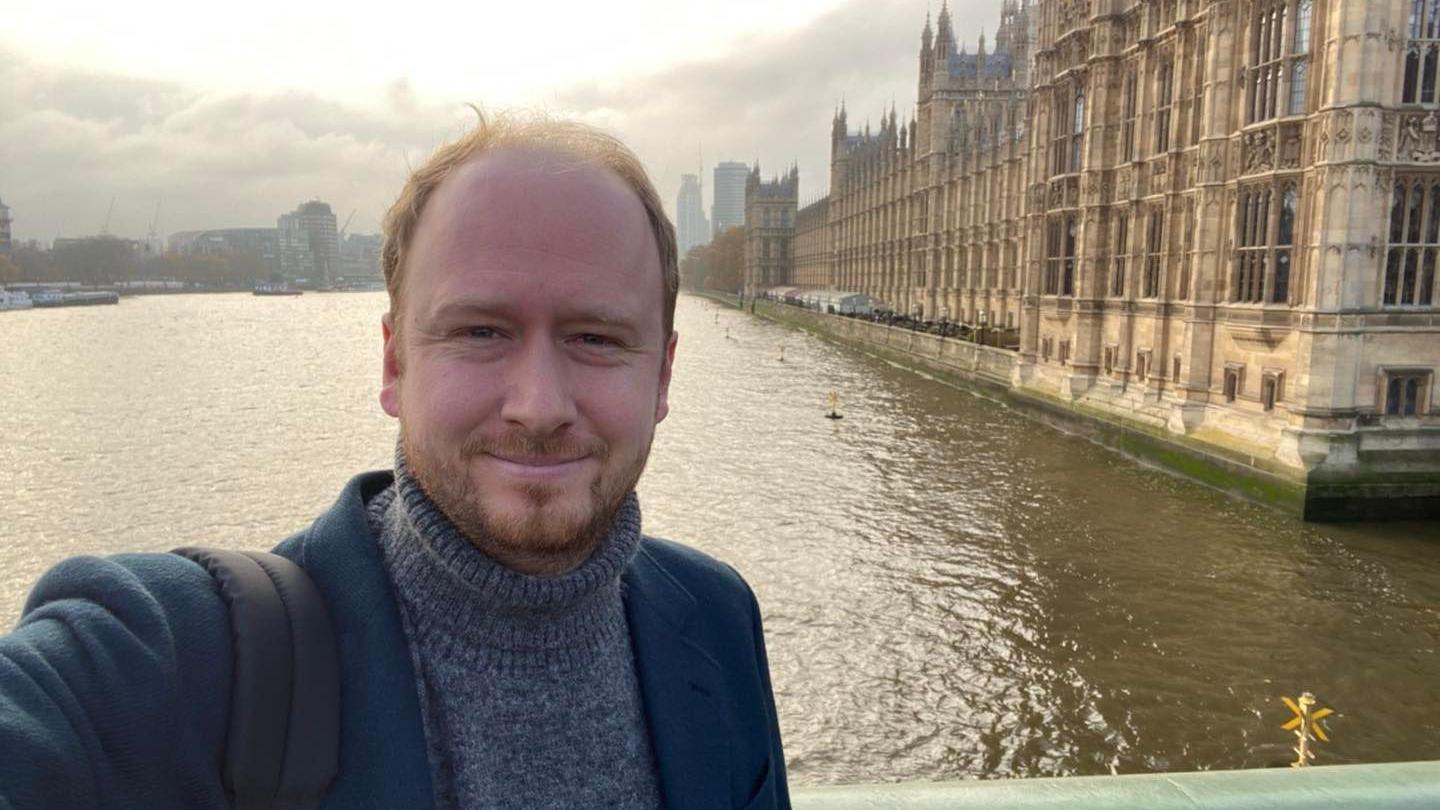 Councillor Tom Briars-Delve wearing a blue blazer and grey turtle neck jumping taking a selfie in front of the River Thames and Houses of Parliament