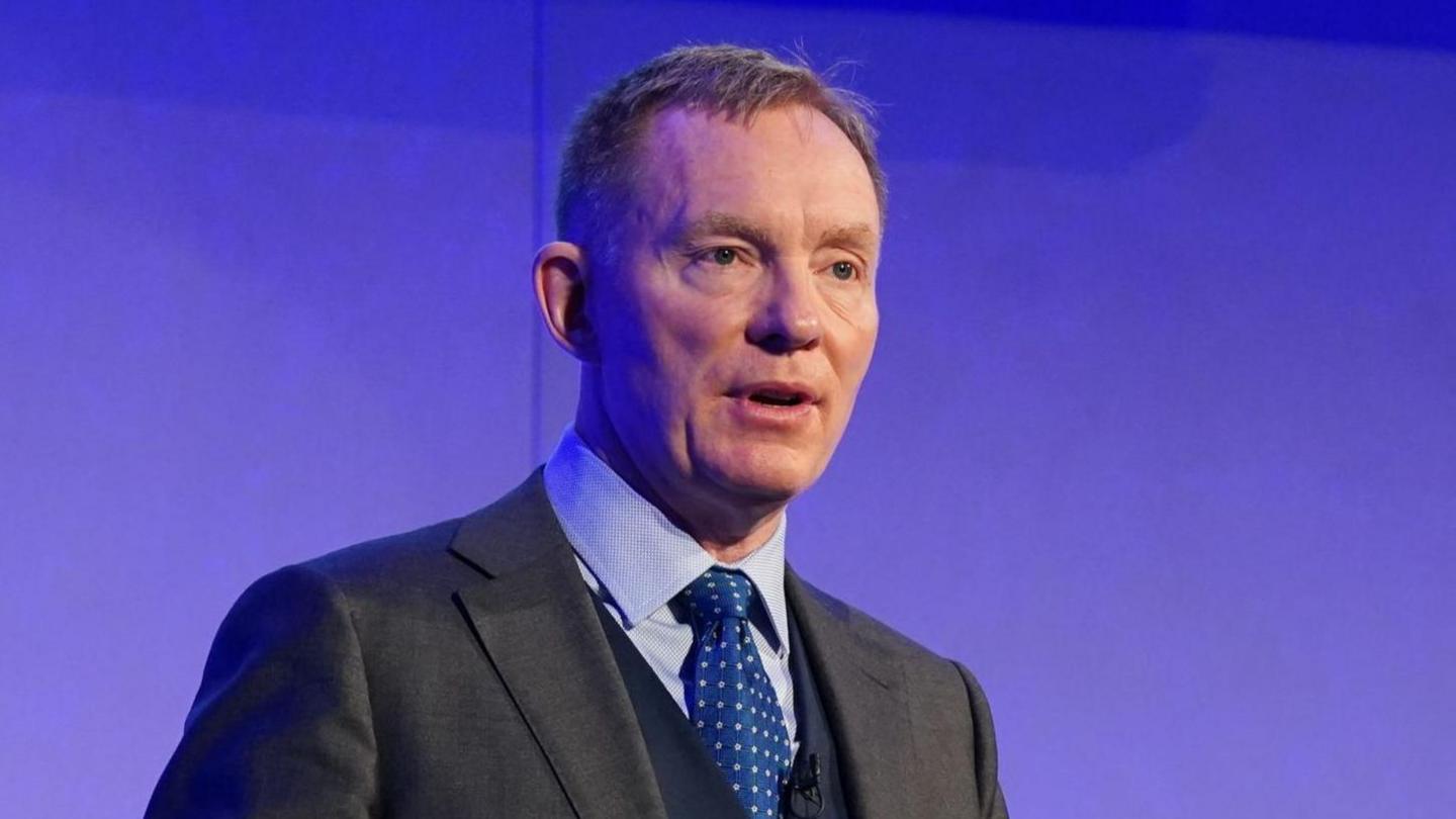 Chris Bryant standing in front of a purple wall. He has a dark grey suit on and a light blue shirt, and blue tie. He is looking to the right of the camera and has short strawberry blonde hair.