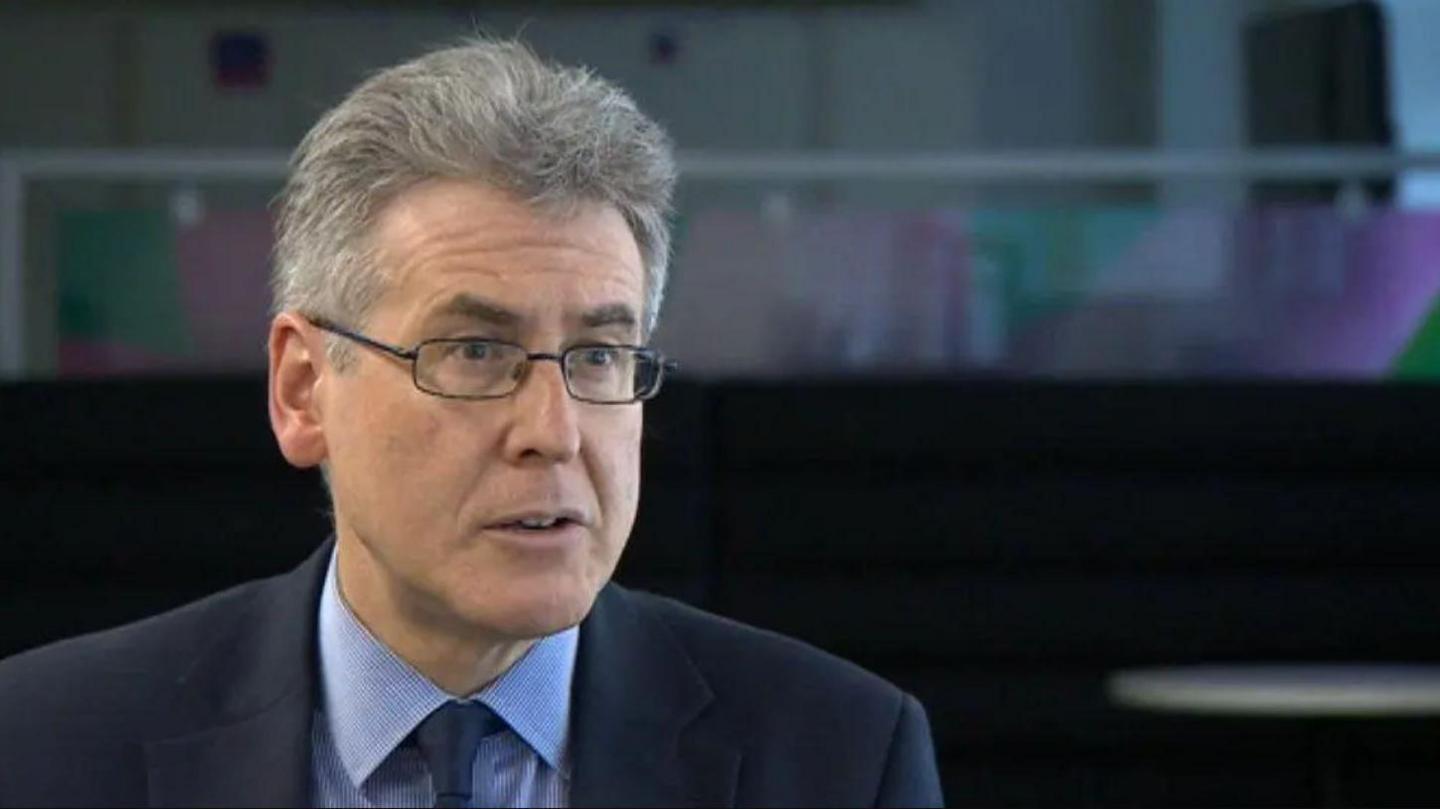 A man with grey hair and glasses in a blue suit and blue shirt against a dark background