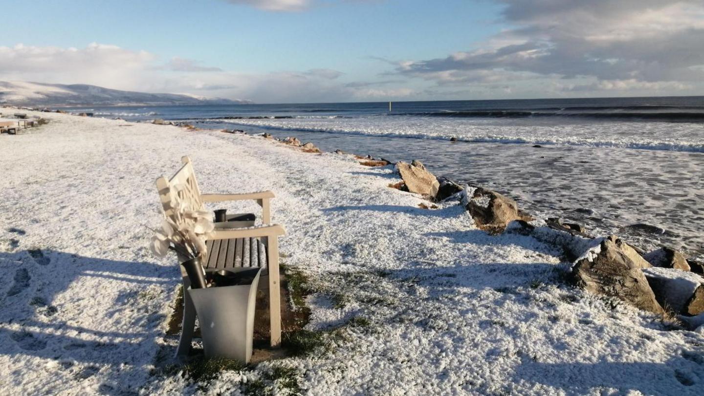 A dusting of snow covers a grassy shoreline. There is a bench and waves rolling in from the sea.