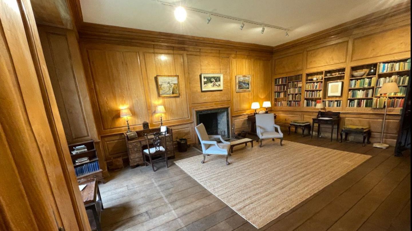 A large wood panelled room has a small desk in it. Two cream armchairs sit by a fireplace. Fitted bookshelves line a wall.