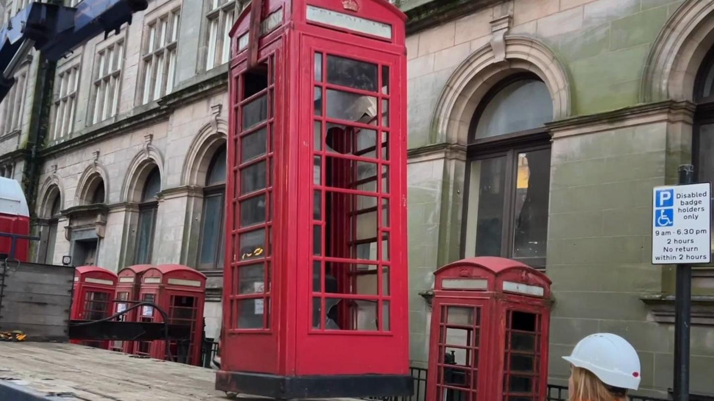 Red phone box on top of lorry and a woman in a hard hat watches on - a row of red boxes can be seen in the distance 