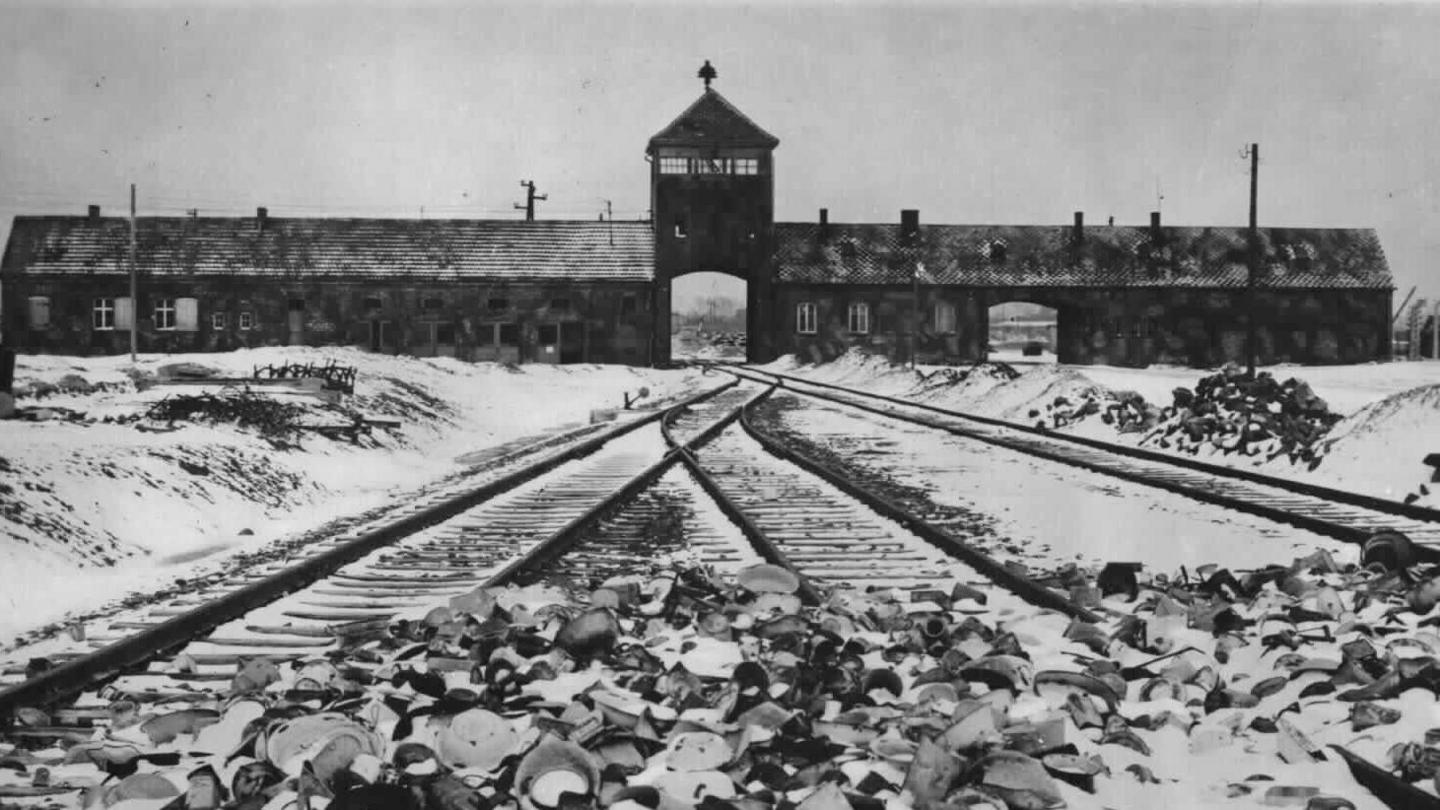 A black and white image of the Auschwitz concentration camp in 1945. Train tracks run up to the entrance of the camp. Snow has fallen on the ground.