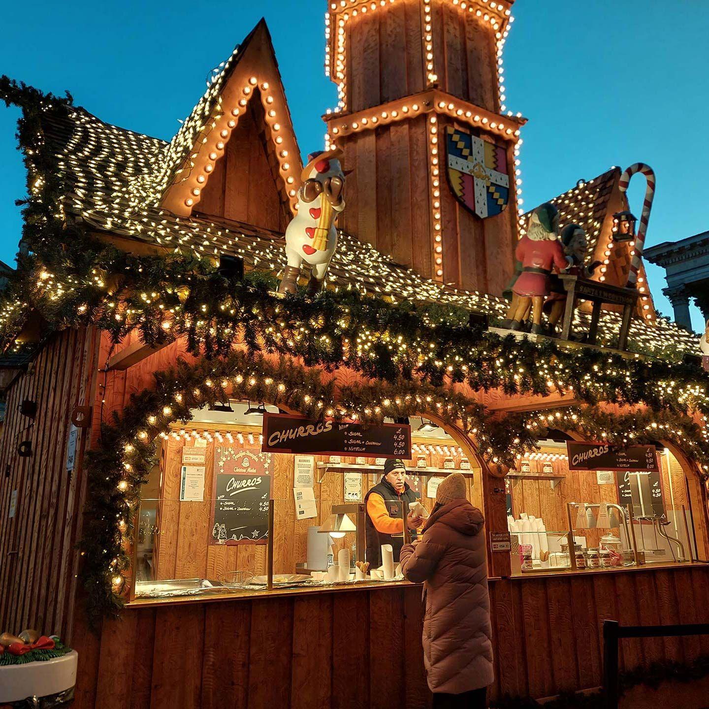 A stall selling churros at a Christmas market. The wooden stall is adorned with fake Christmas tree sprigs, golden fairylights, signs saying "churros". A man stands behind the counter at the stall, serving a woman in a long winter coat