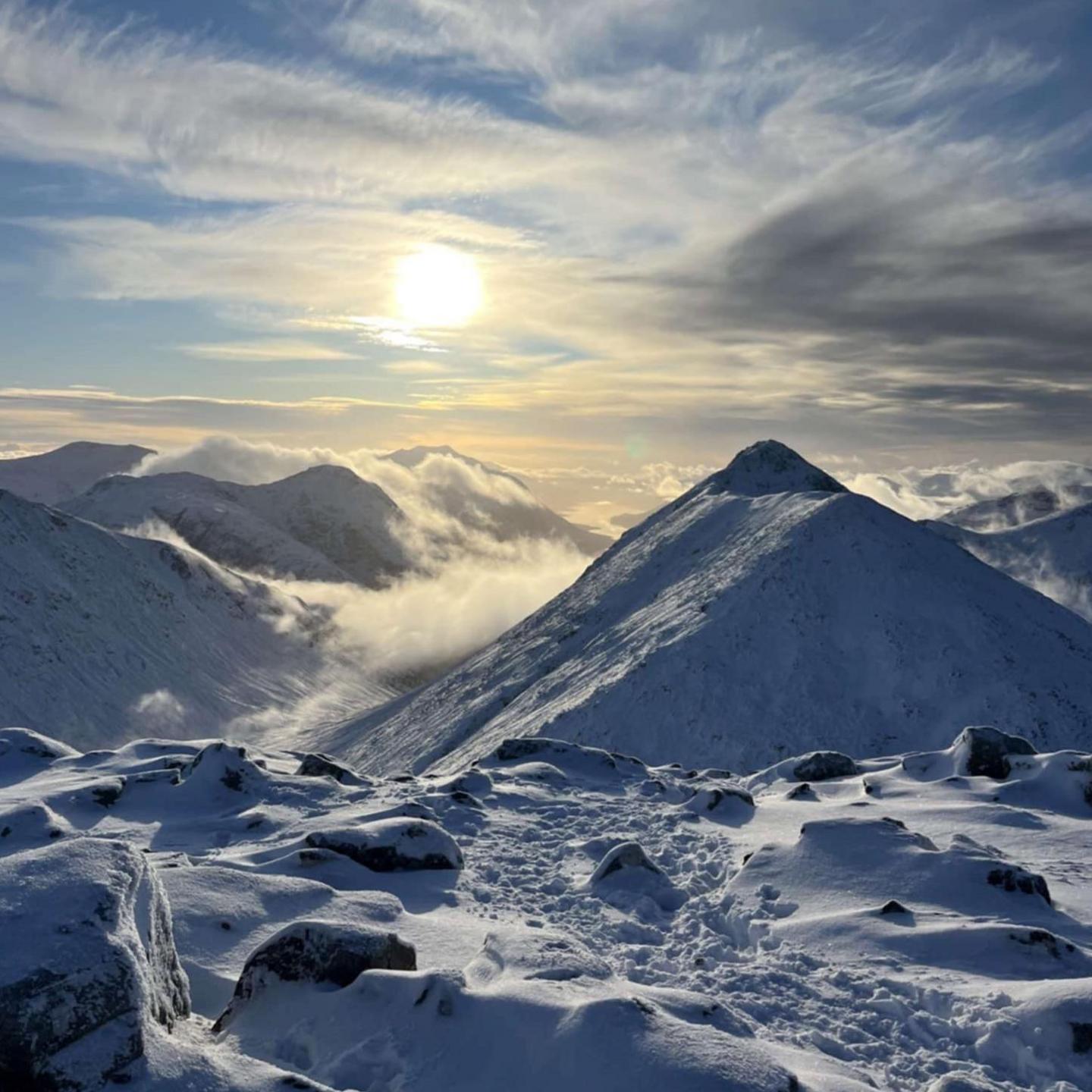 The sun shines behind clouds above a snowy mountain landscape. Low cloud hangs in glens between the mountains.