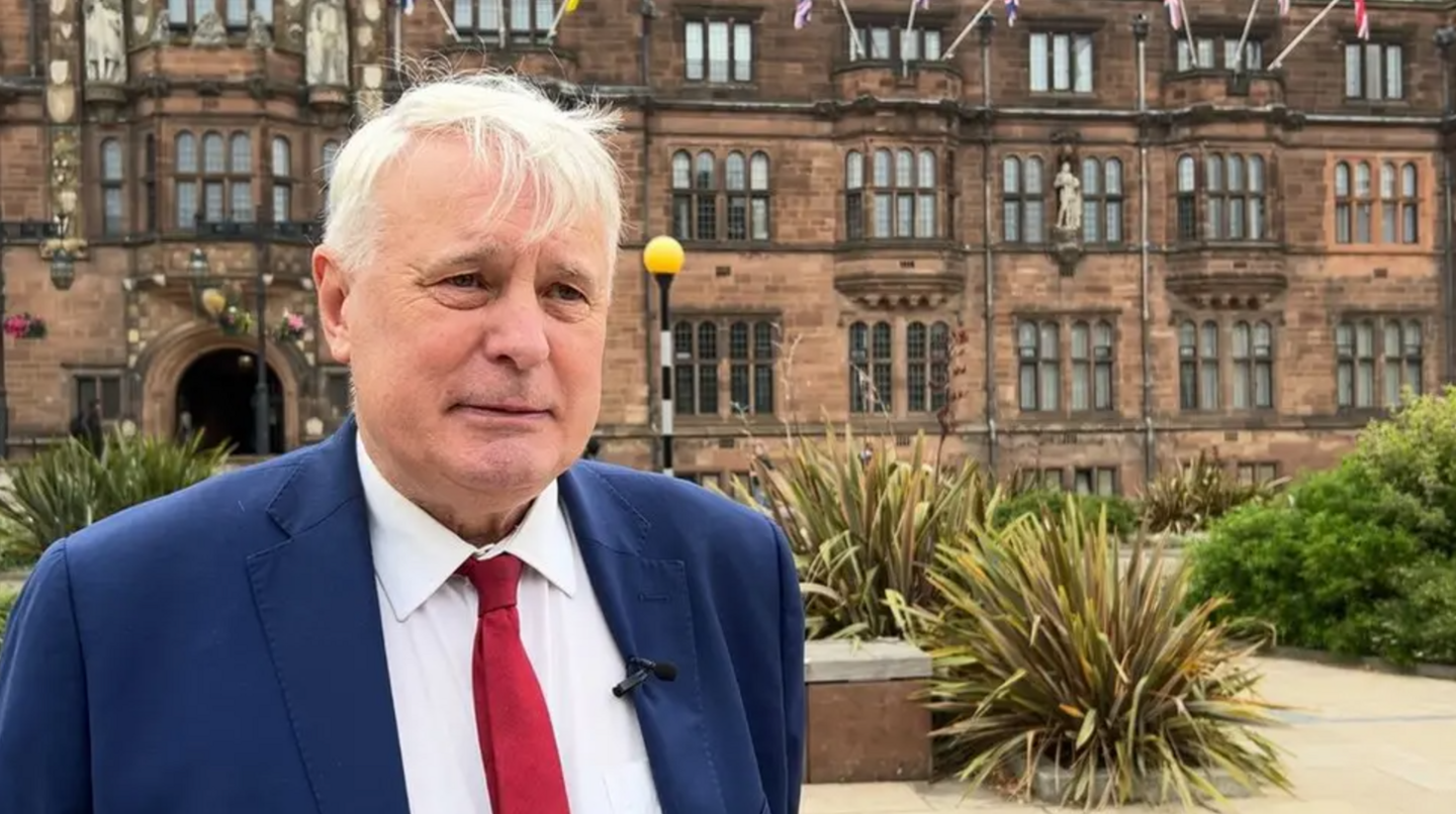 A man with white hair in a blue suit jacket, white shirt and red tie, stood in front of a large red brick building with bushes behind him