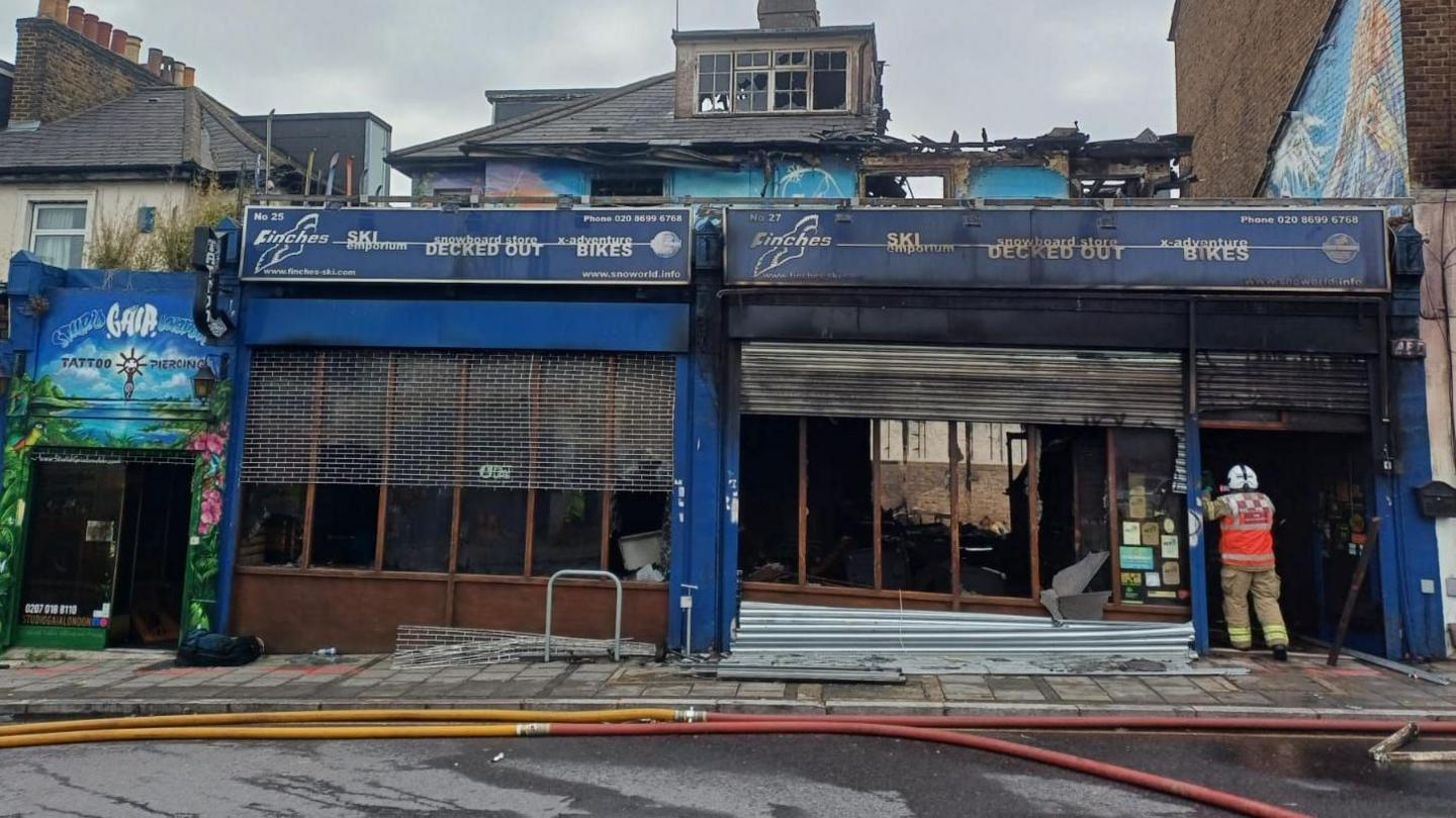 A red brick building with a shop front and signage in dark blue, which is burnt out inside