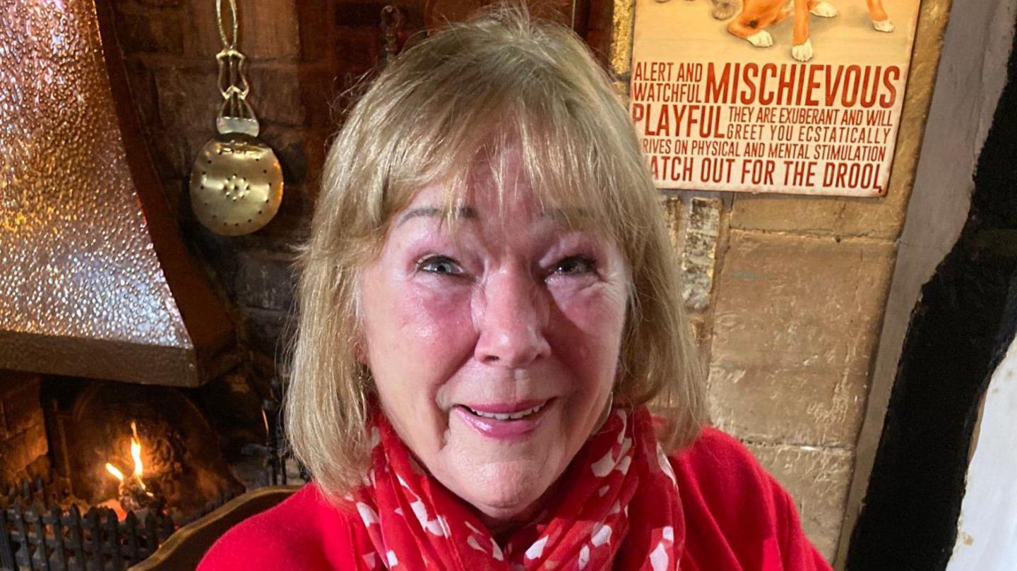 Sheila Williams wearing a red top and scarf sitting in a pub by the fireplace. She is smiling and has shoulder-length blonde hair.