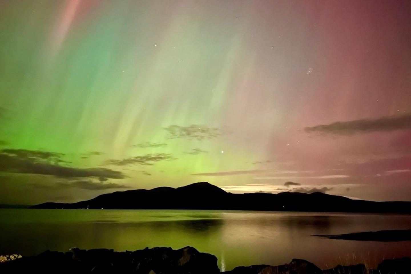 A green and pink aurora over hills and water.