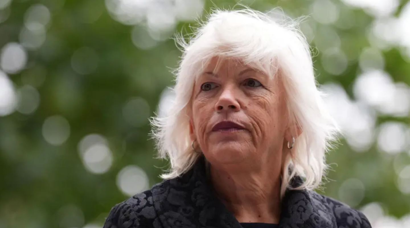 Women with white hair looking to the side of the camera, upwards. She is wearing a black coat. In the background the leaves of a tree, out of focus.