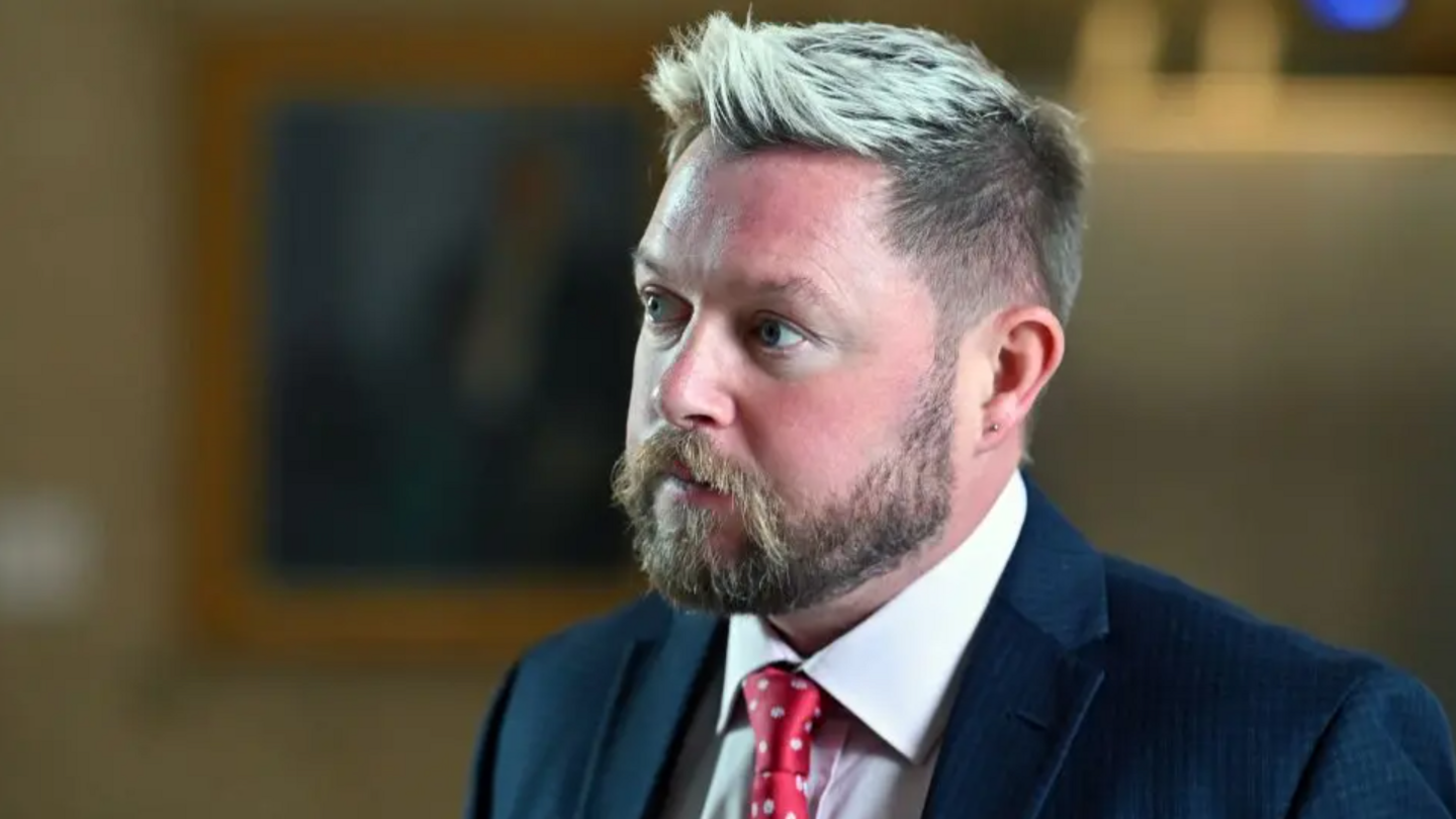 Jamie Greene looking slightly off camera. He has partially dyed blonde hair which is spiked up at the front. He has a long, light-coloured beard. He is visible from the shoulders up. He is wearing a blue suit over a white shirt with a red tie which has a white polka dot pattern.