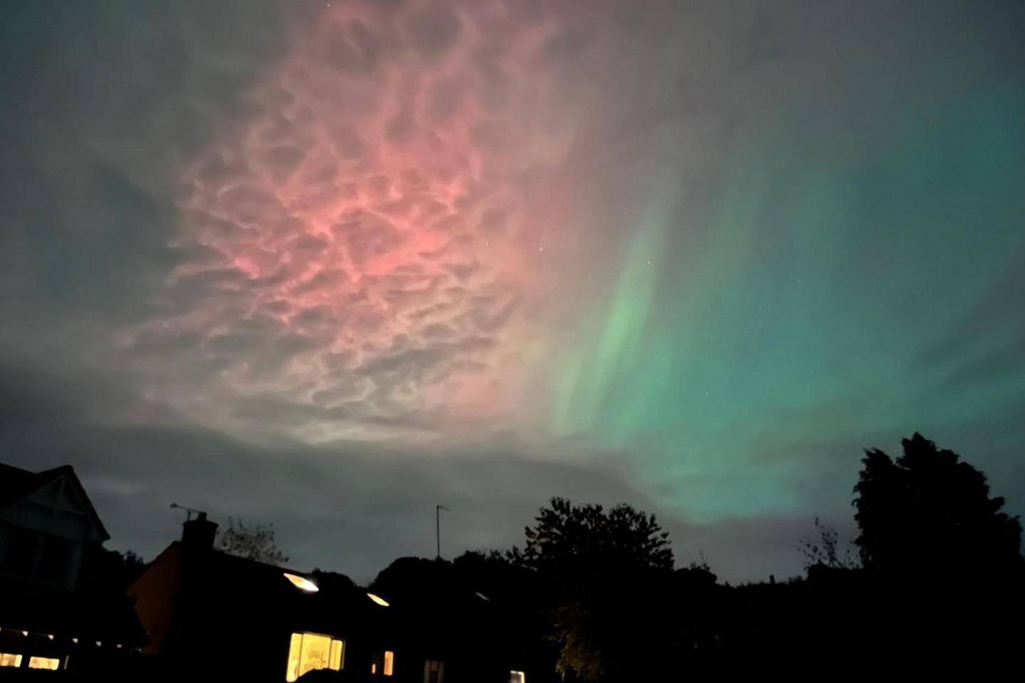 The Northern Lights appear in pink and green behind clouds and above a house next to trees.