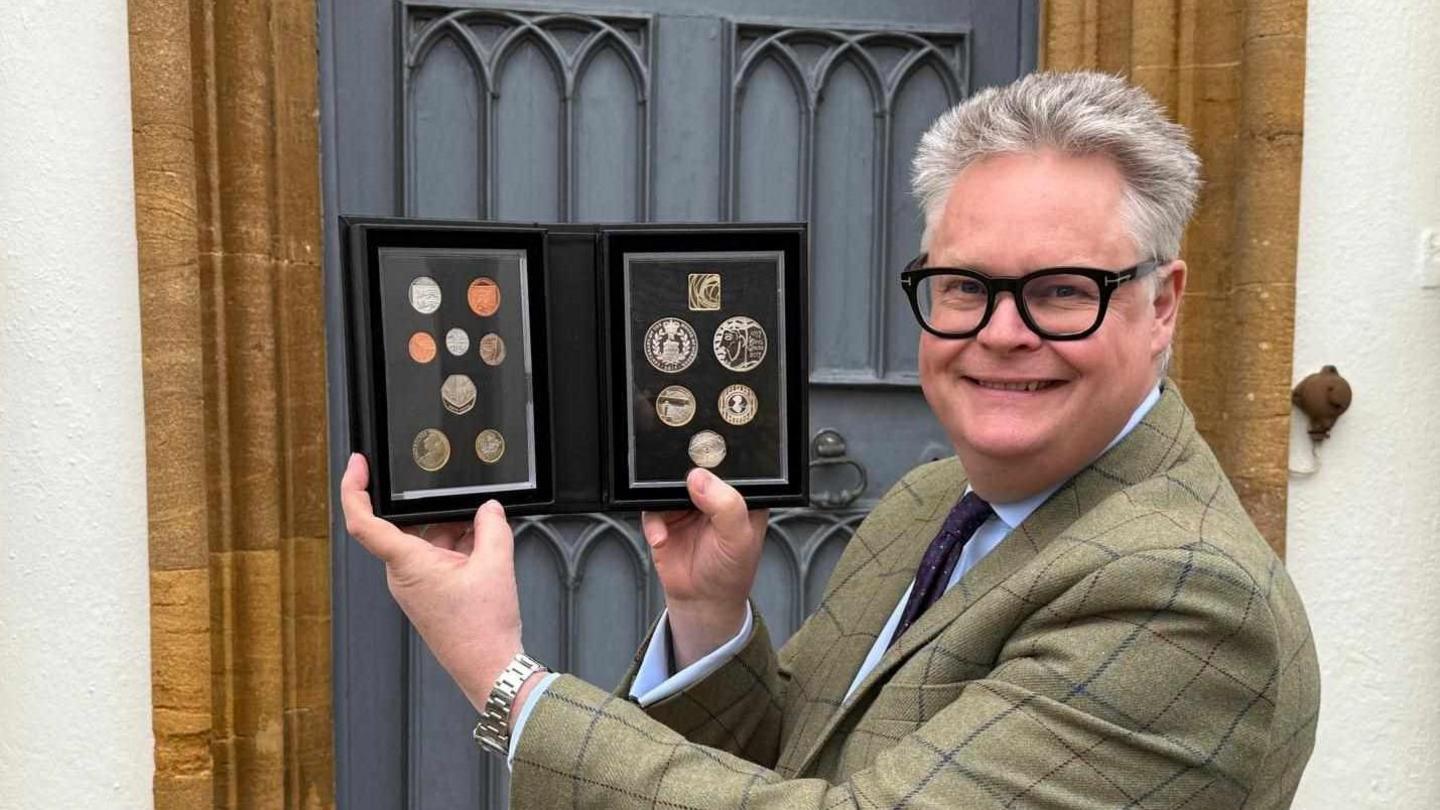 Auctioneer Richard Bromell smiling at the camera, wearing black-framed glasses and a tweed jacket, pictured holding a coin collection.