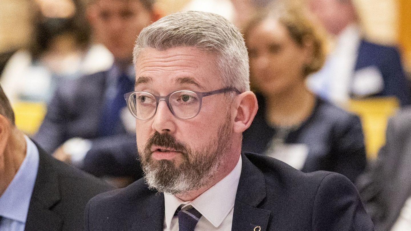 Niall Ó Donnghaile has grey hair and grey glasses.
He also has a beard that is predominantly grey. 
He is wearing a navy jacket, white shirt and navy tie.