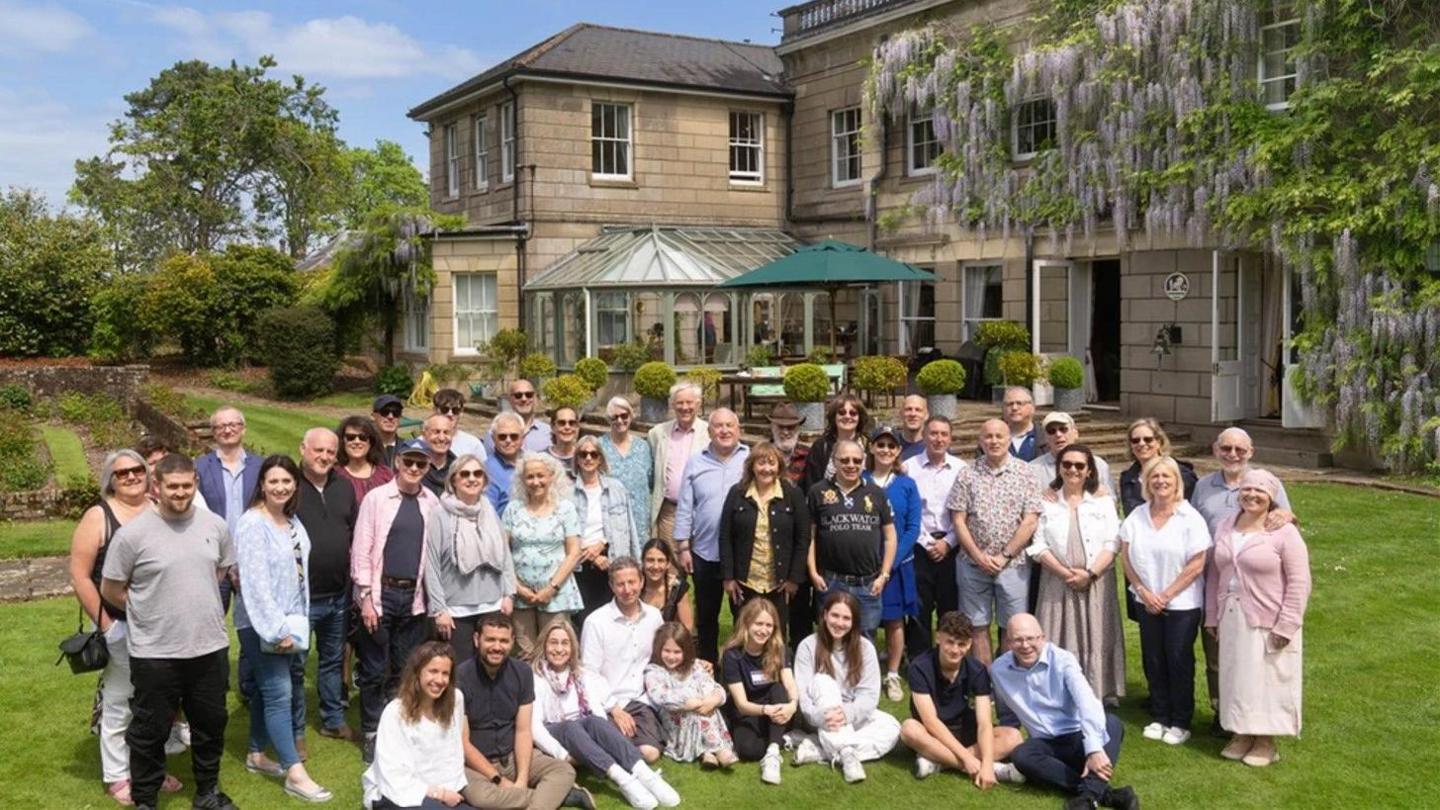 A group of about 50 people posing for the camera on the lawn outside Wintershill Hall - a large stately home with flowering wisteria covering the walls.