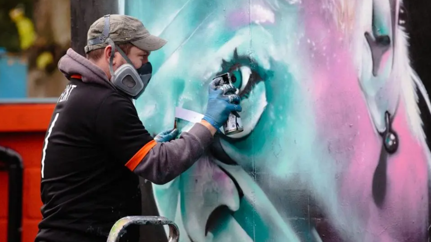 David Nash, wearing a face mask, spraying his Keith Flint mural on to a water tank at Braintree Town FC.