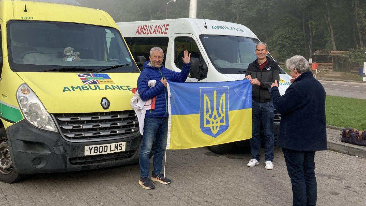 Two men, holding a flag, standing between two ambulances, with a person standing in front of them putting their thumb up. They are all smiling. 