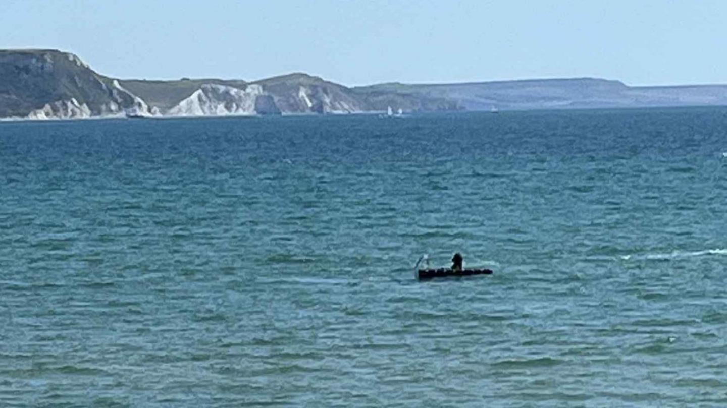 A blue sea that has a black swim raft on. There are grey and green mountains in the background of light blue sky. 