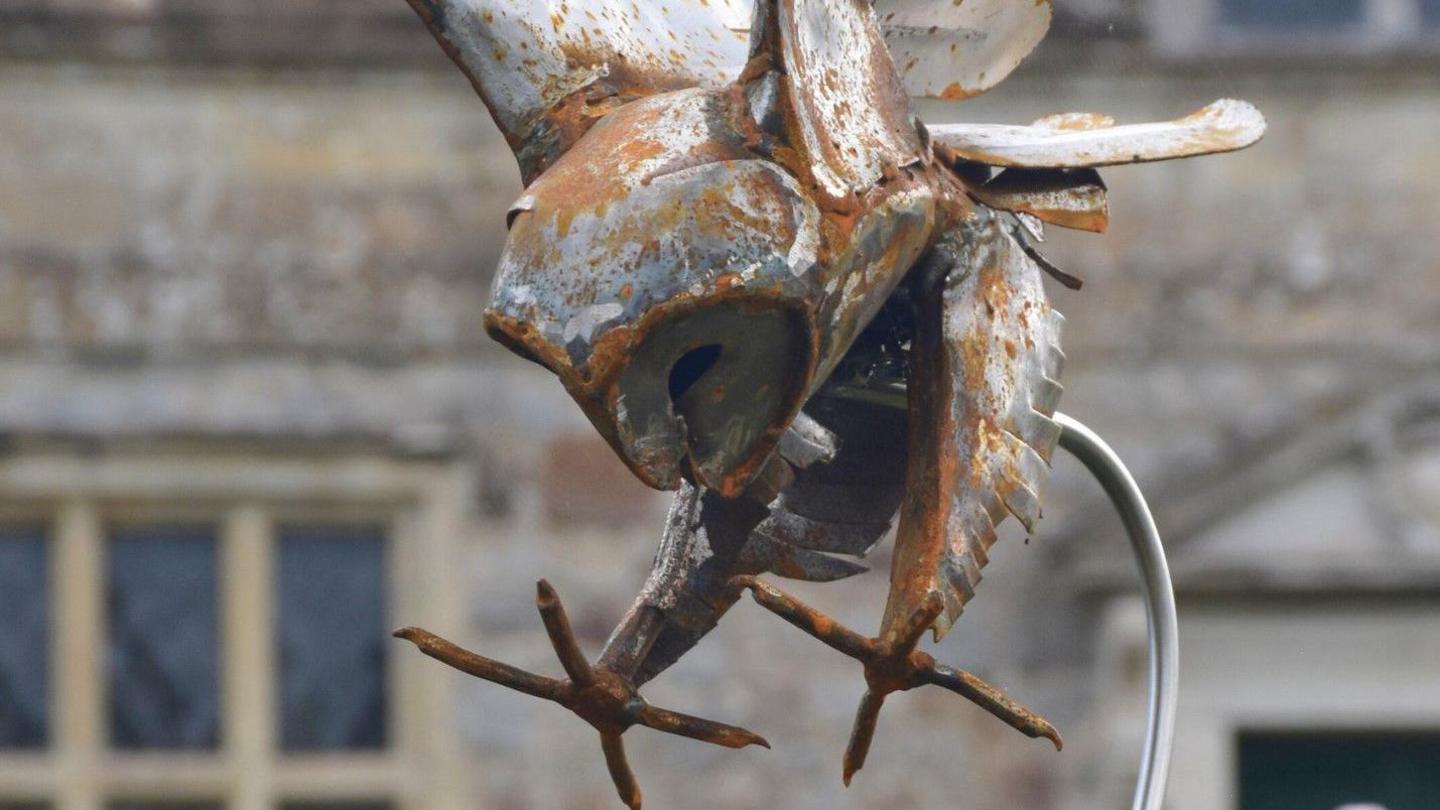 A rusting metal owl captured in flight, head down and tallons out, looks to be about to catch prey in Swooping Owl by Andy's Metal Art. The windows of Avebury Manor are visible behind it. 