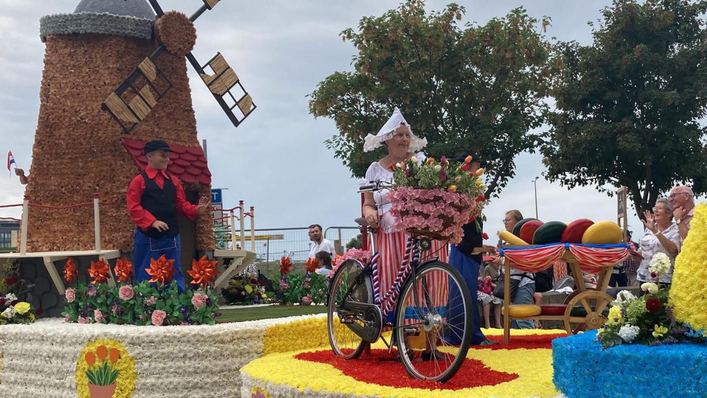 A series of floats used in the Battle of Flowers earlier this year. One has a windmill and someone dressed in a black cap, and red shirt. The other has someone dressed in a white hat, top, and red and a red and white striped dress, standing beside a bicycle full of flowers.