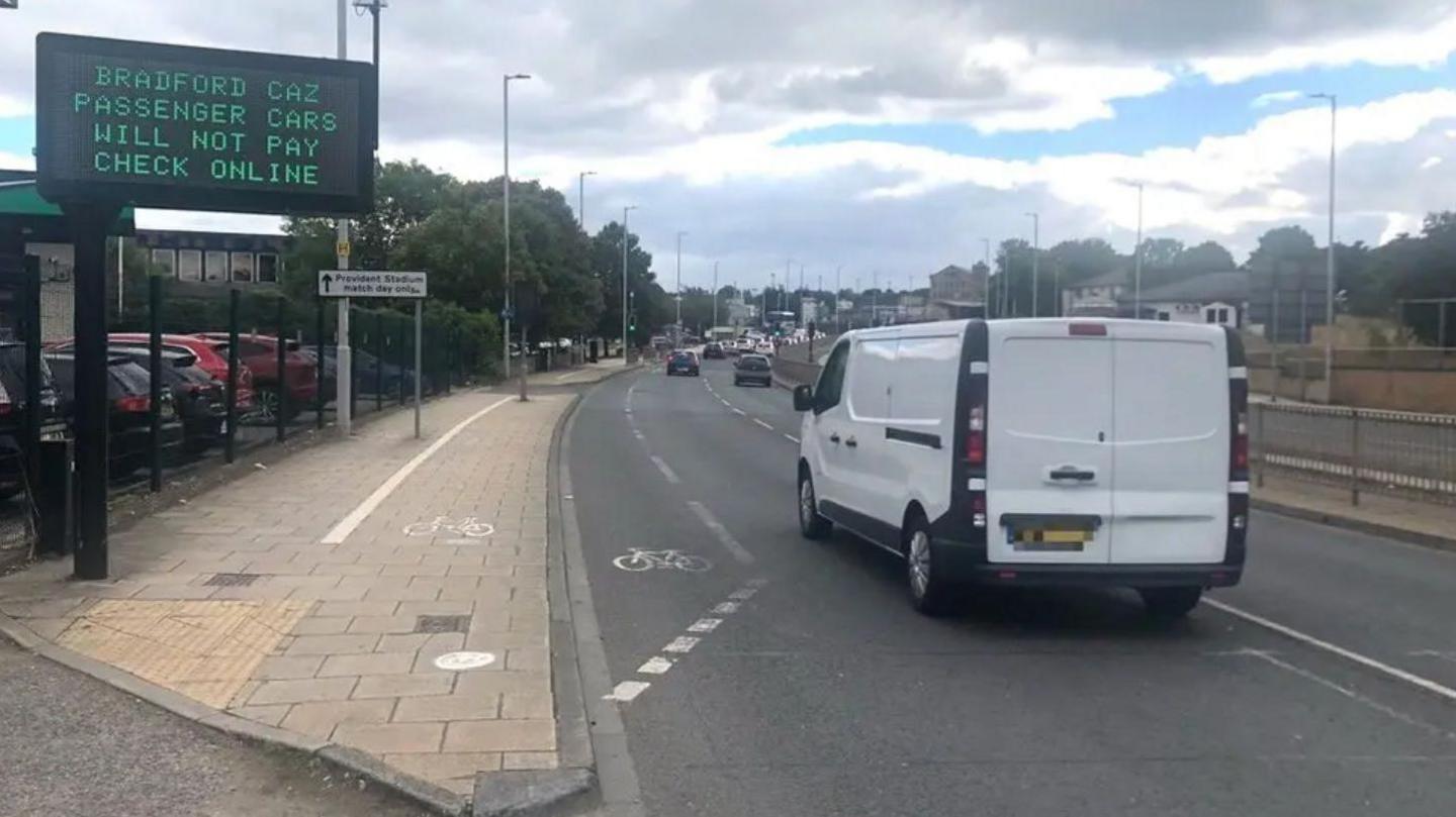 An image of a road shows a white van travelling along it with a sign on the pavement on the left. It reads "Bradford CAZ passenger cars will not pay check online".