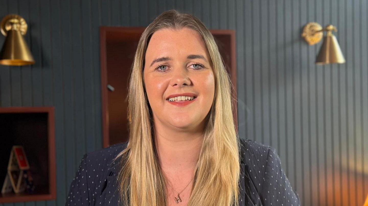 Heather Stewart has blonde hair to below her shoulders. She is wearing a navy jacket with a patter. She is also wearing a small chain with the letter H on it. The background is a green panelled wall with two gold lights and wooden frames hanging.
