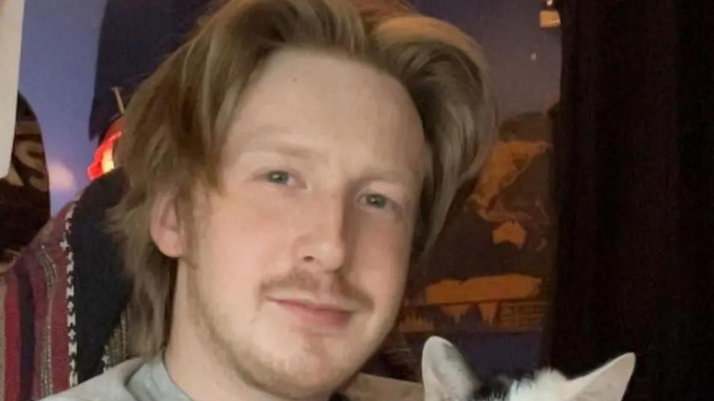 Mr Smith has long blond hair, parted in the middle and facial hair. He appears to be holding a black and white kitten, although only the ears of the animal can be seen.