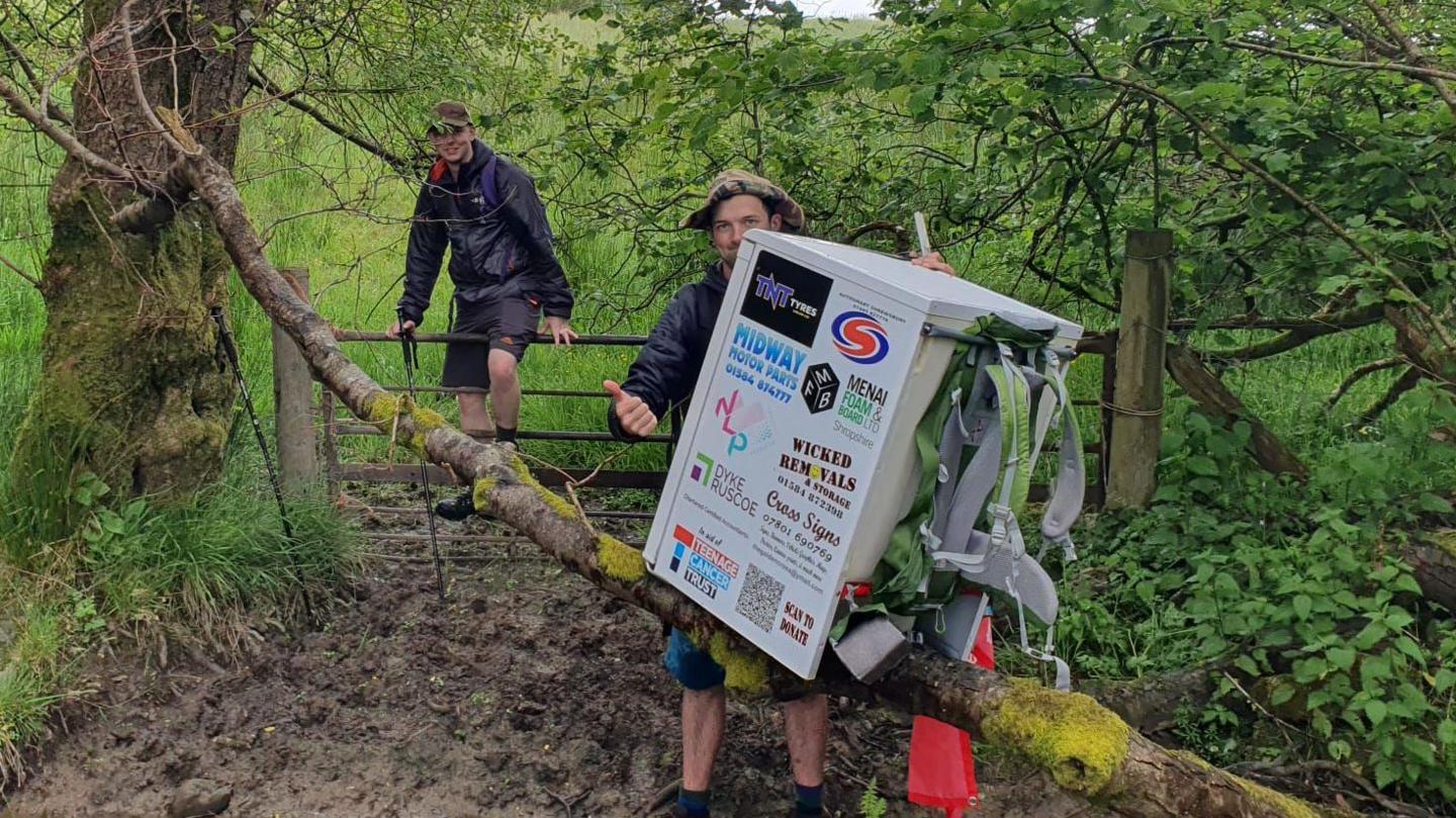 a man balancing a fridge on a tree branch next to a brook