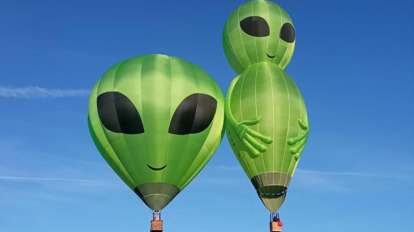 Two green hot air balloons flying in the sky with alien faces and small brown baskets beneath them. One balloon is round, the other is taller and has hands with four fingers.