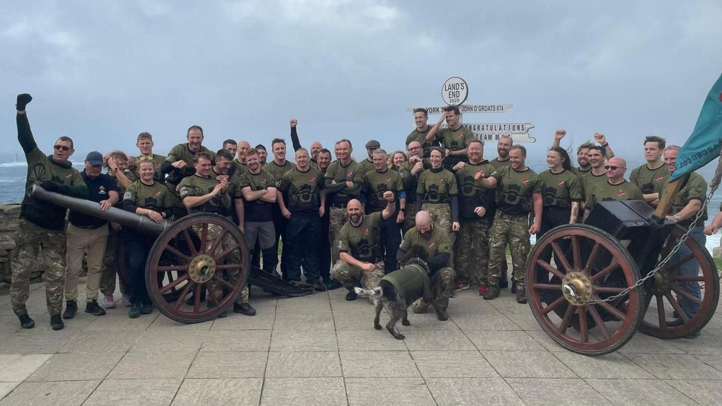 A of 37 people stood in three rows. There is a sign for Lands End in the background and the sea can be seen to the right of the picture. They are dressed in khaki and one person kneeling at the front is fussing a black and white dog which is wearing a khaki coat. To the left and right of the photo are the wheels and gun which they have dragged along the route