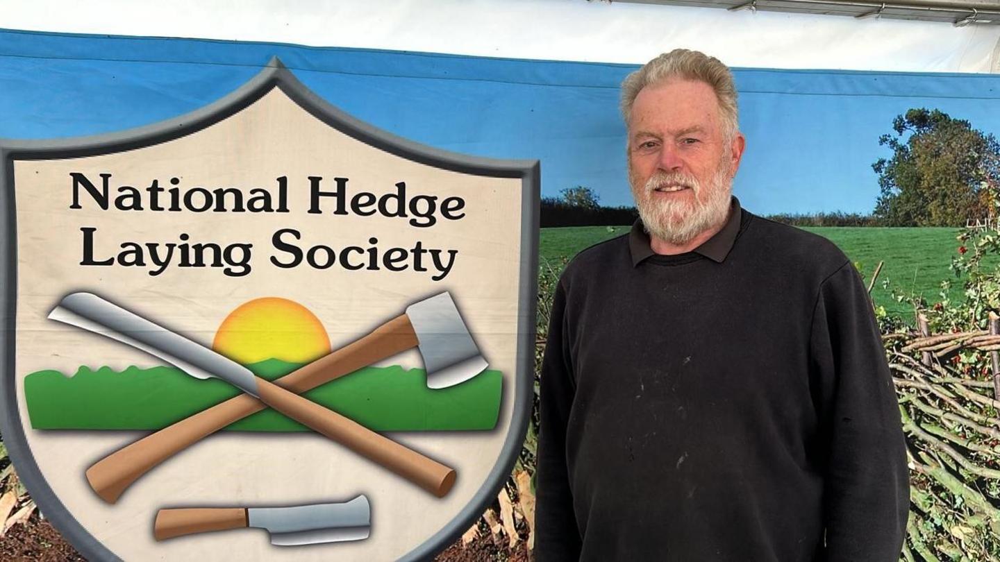 Stephen Budding has white hair and a beard. He is wearing a black jumper. Mr Budding is standing in front of a banner depicting the National Hedgelaying Society logo which depicts an axe and other hand tools.