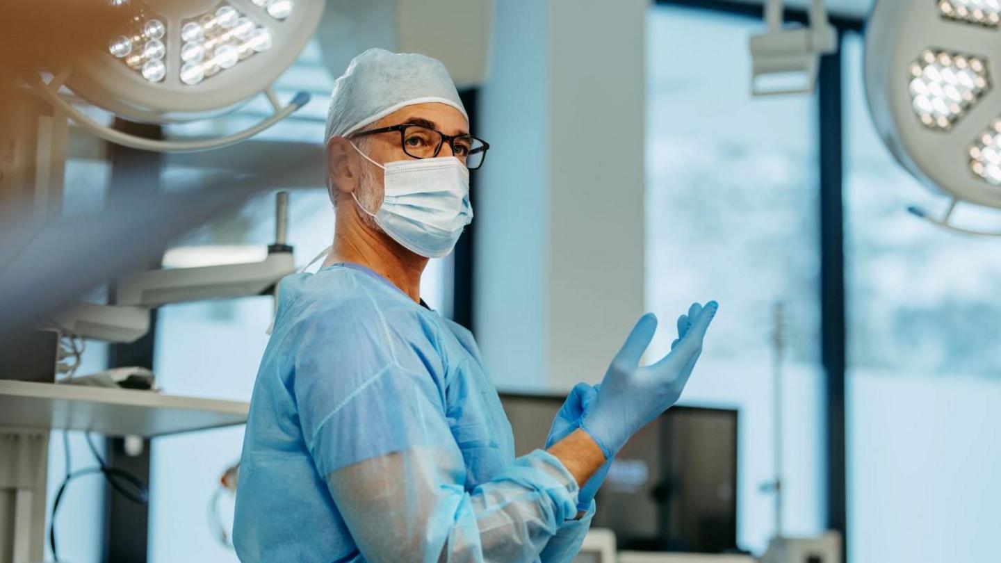 a man in glasses, a hairnet and surgical scrubs pulling a glove onto his hand, in a hospital room setting