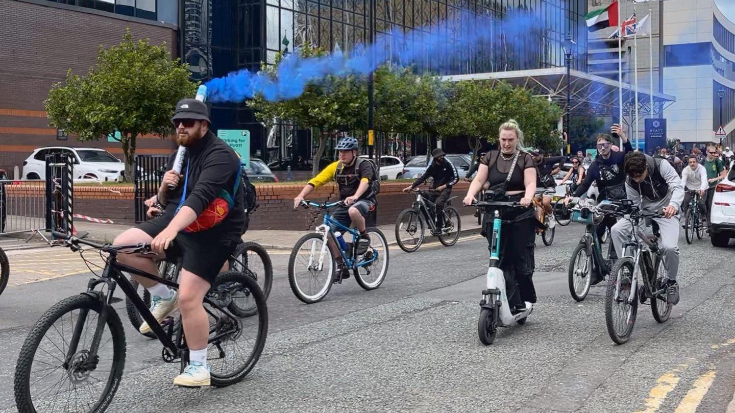 A man is wearing a black top and shorts and is cycling on a bike. He is holding a smoke flare that is letting out blue smoke into the air. People are following him on bikes and scooters