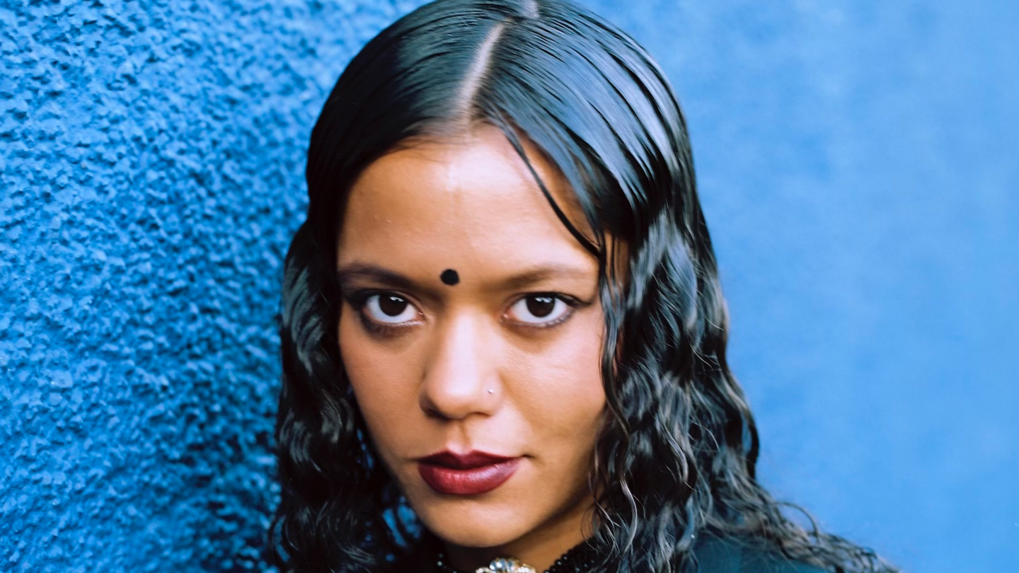 A woman with long black hair and red lipstick looks towards the camera, against a blue background