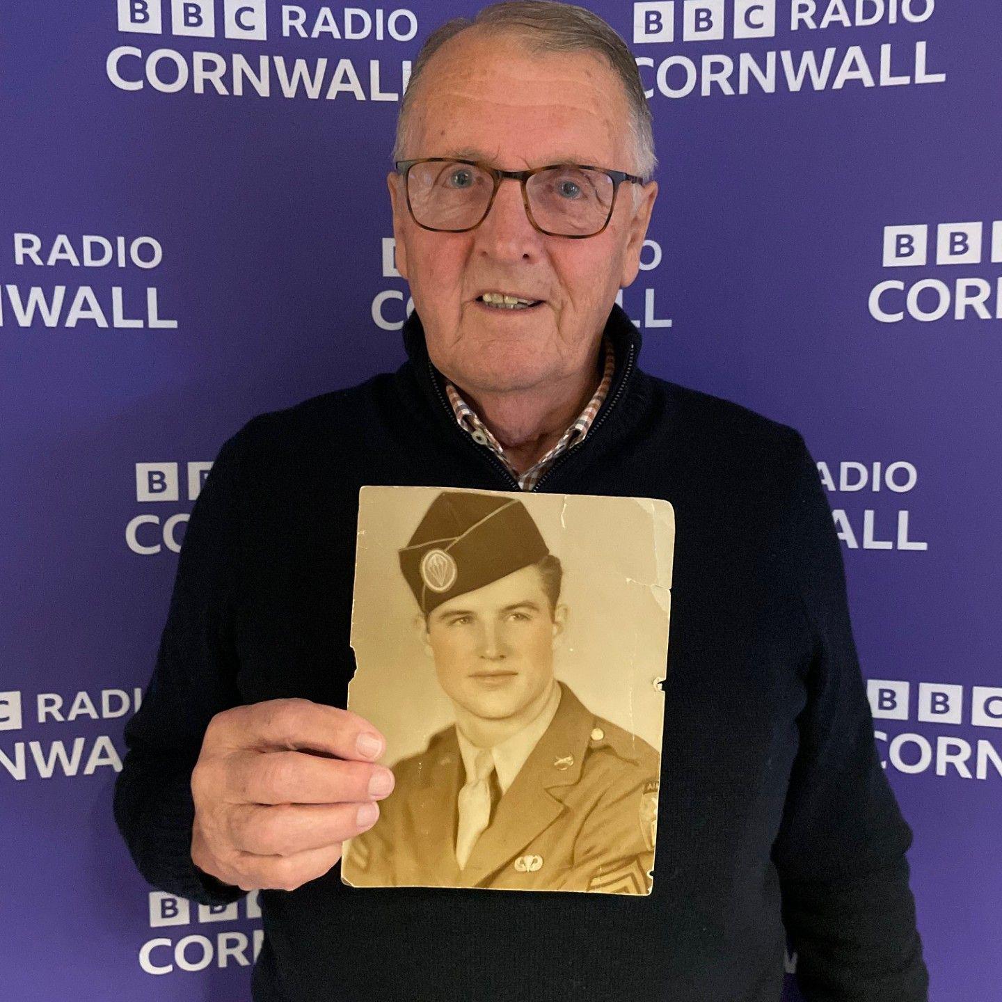 Tim Lobb is wearing glasses, a black jumper and collared shirt and is holding an old photograph of his father. He is standing in front of a BBC Radio Cornwall screen.