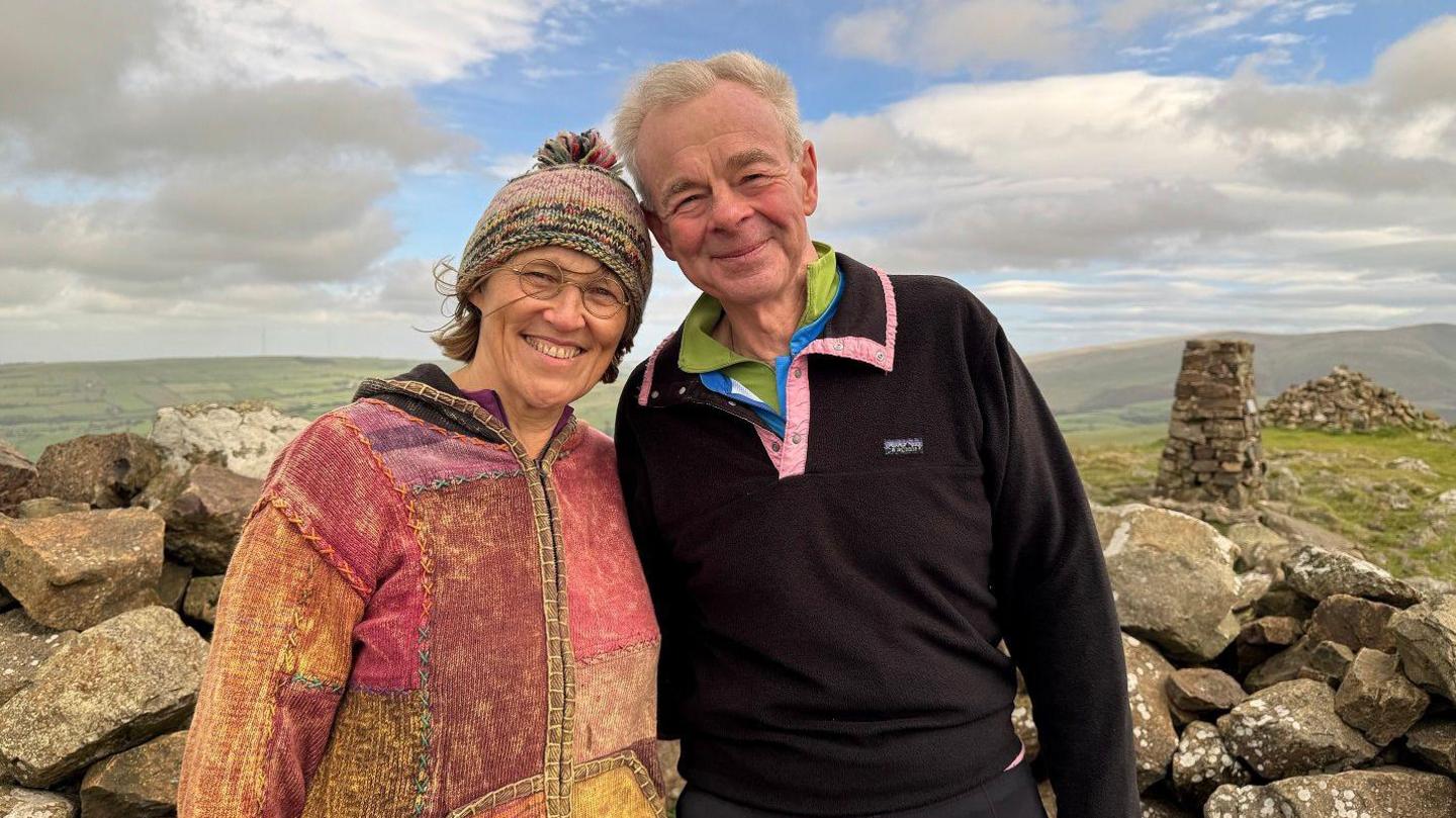Catherine Wetherfield and Ian Teasdale standing on a hilltop. Catherine wears a wooly hat and jacket that is pink patchwork. She had short brown hair and wears round-shaped glasses. Ian has short white hair and wears a black fleece with a pink trim. They are both smiling. There are large stone and the remains of stone buildings behind them. The tops of hills are in the background. 