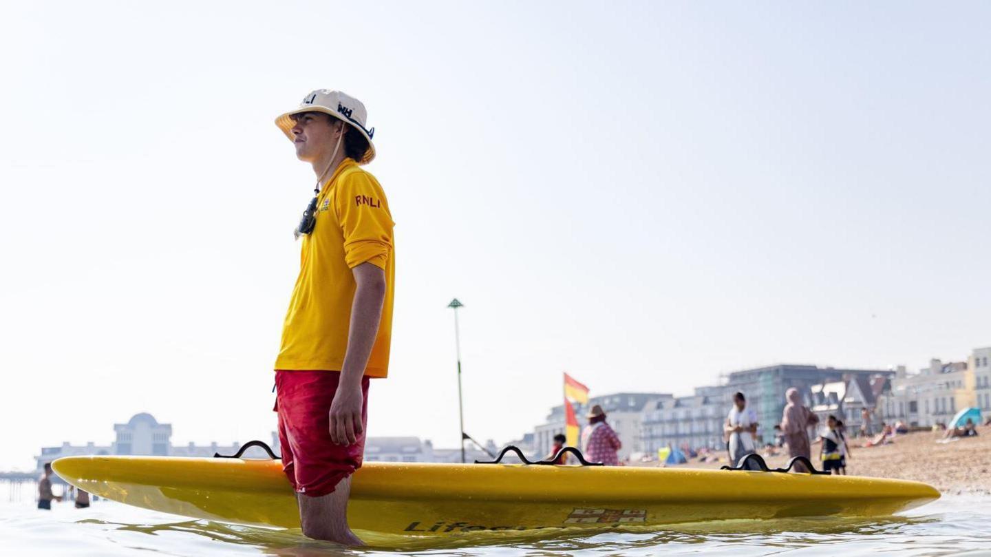 A lifeguard stands in the water next to a bright yellow paddleboard keeping watch at Southsea beach in Portsmouth. They are wearing red long shorts, a yellow short sleeved polo shirt and a yellow bucket hat