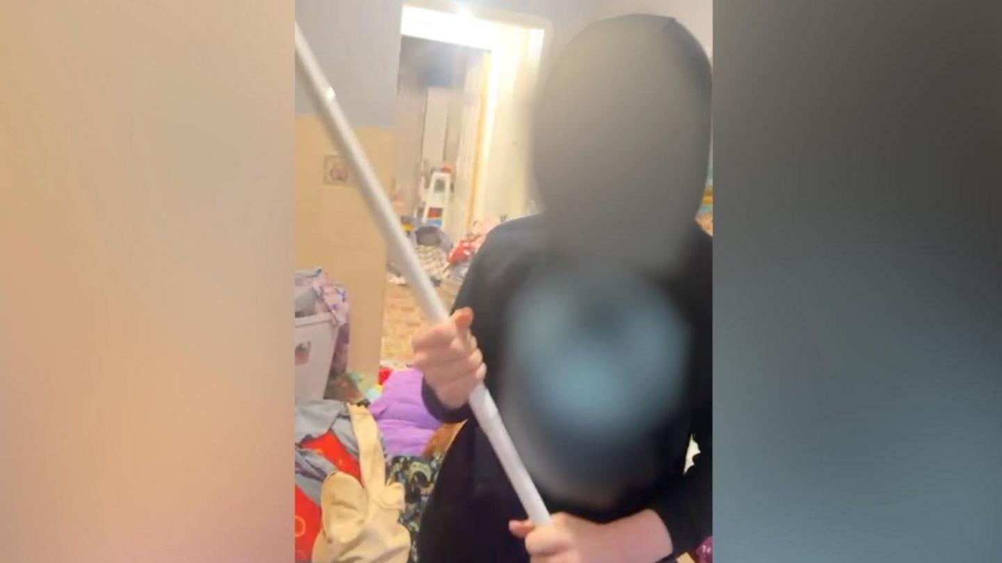 A still image, taken from CCTV footage, of a teenage boy with his hood up, threatening his mother with a pole in their living room. His face is blurred to protect the family's identity.
