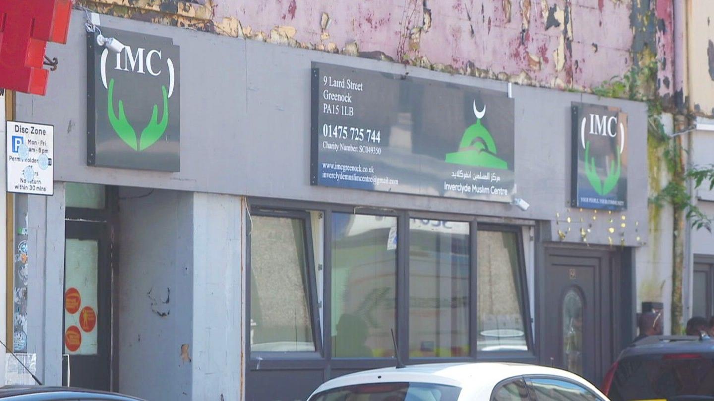 Exterior of a grey building with black doors and windows and peeling paint on the first storey. Above the entrance is a sign saying "IMC" for Inverclyde Muslim Centre.